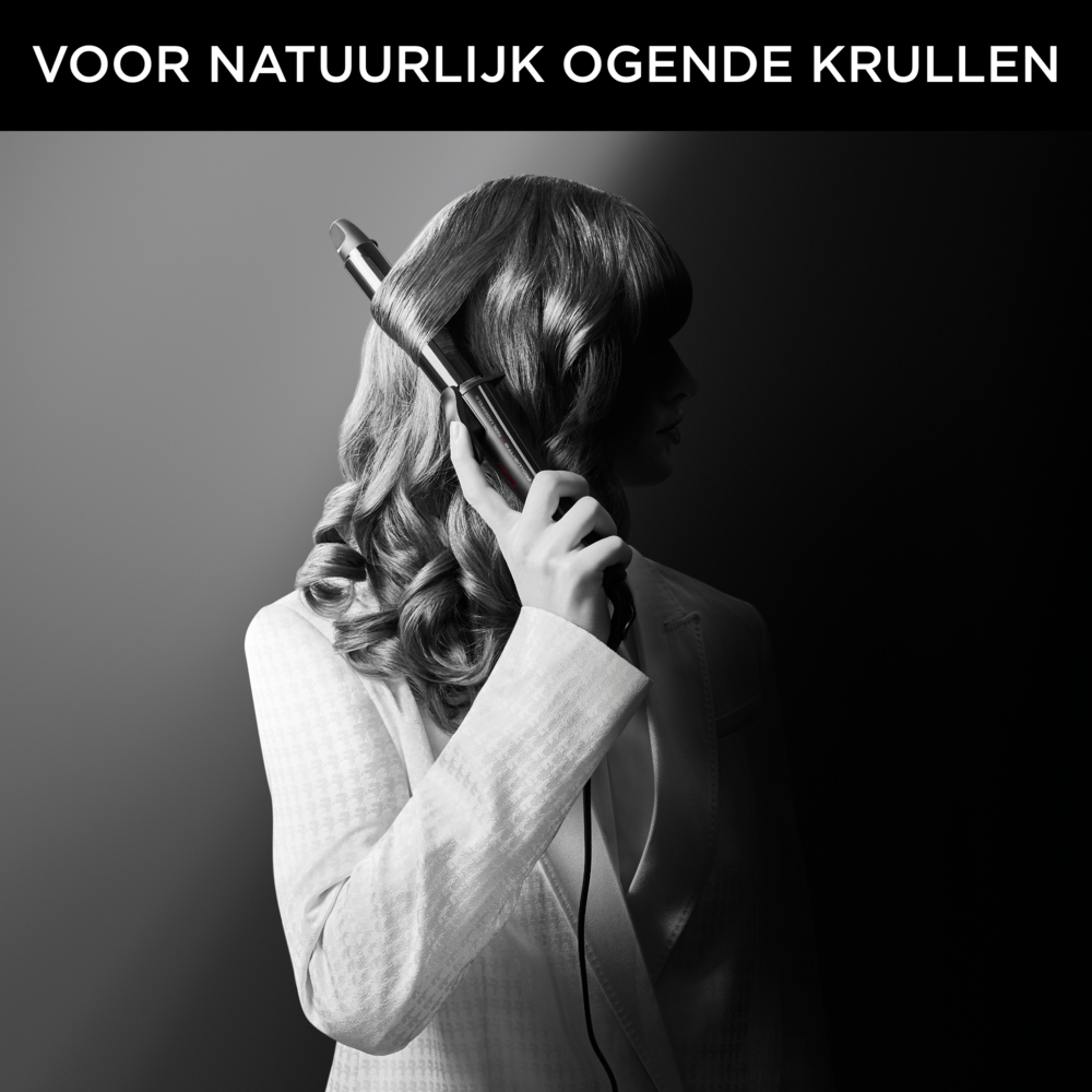 Kobieta kręci włosy lokówką. Ma na sobie białą kurtkę. Tło jest ciemne.