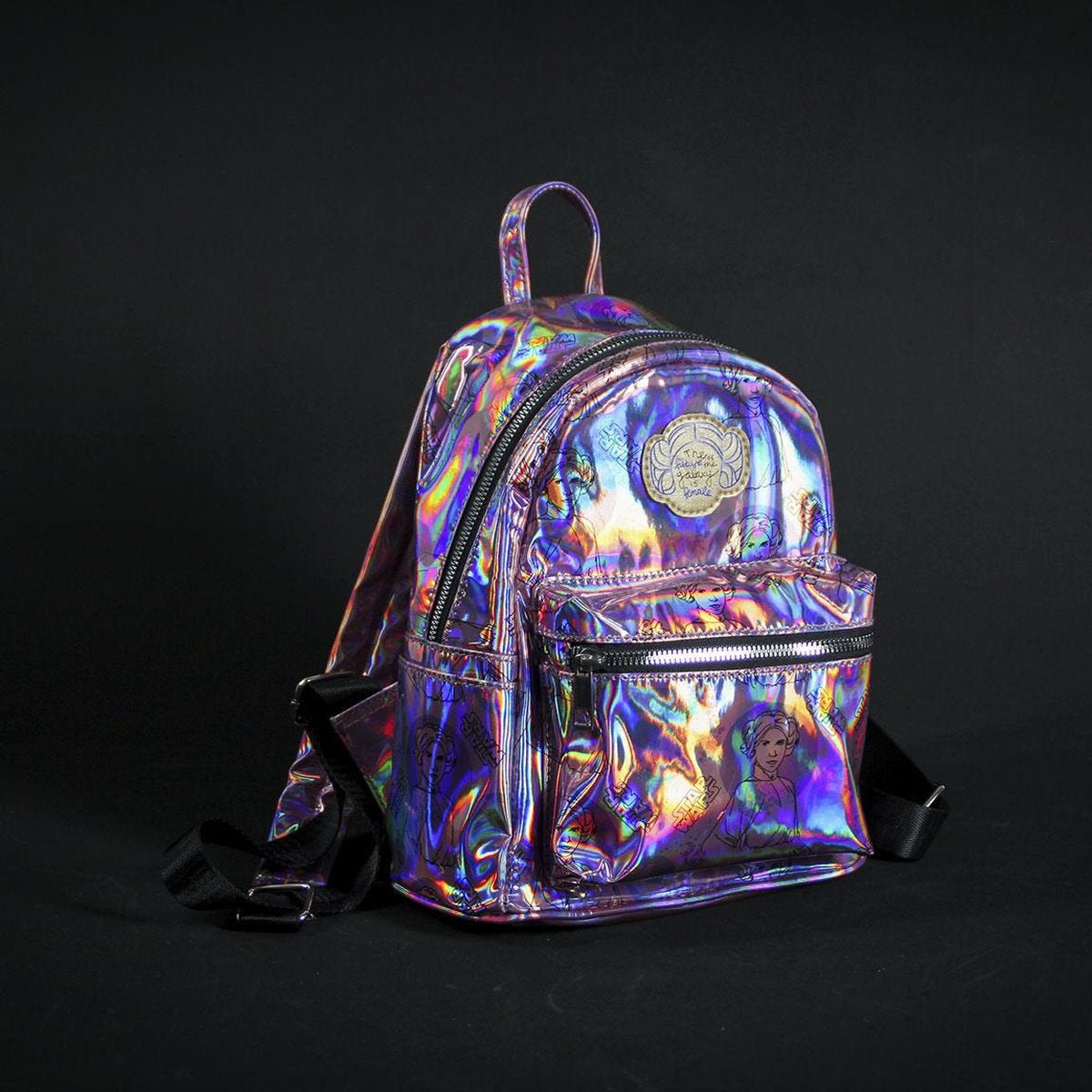 Una mochila brillante e iridiscente con una correa negra. Incluye un bolsillo frontal y un asa pequeña, sobre un fondo negro.