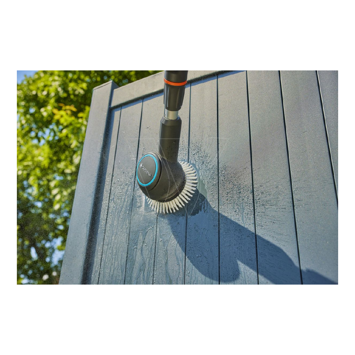 L'acqua viene spruzzata da una spazzola nera su una staccionata di legno grigio. La spazzola ha dettagli blu e un tubo collegato.