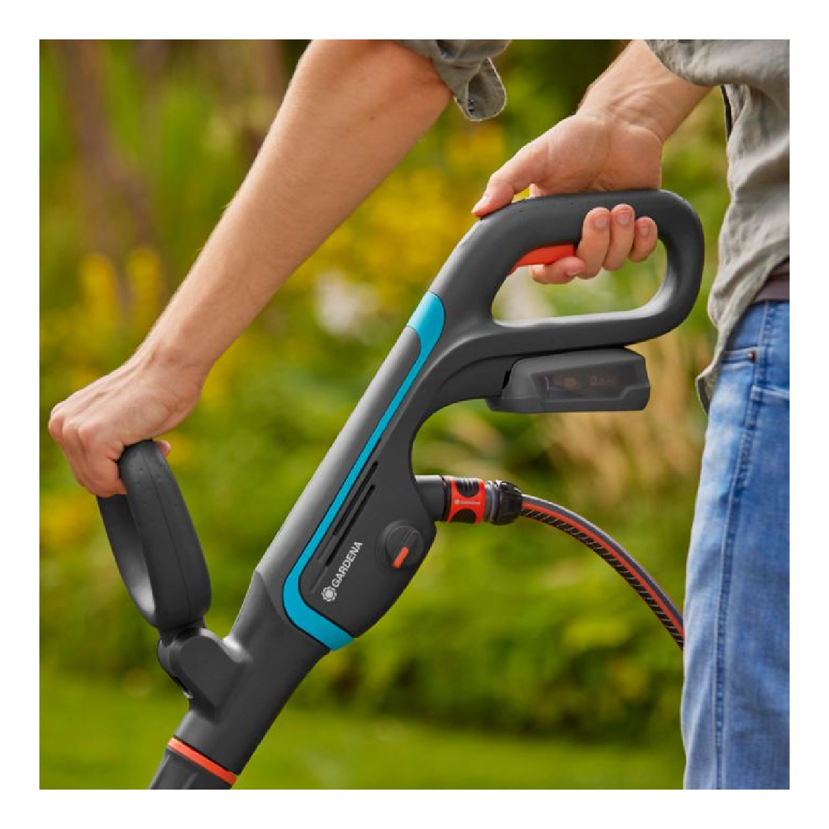 Eine Person hält den Griff eines Reinigungsgeräts. Der Griff ist schwarz mit blauen und orangefarbenen Details. Blaue Jeans sichtbar.