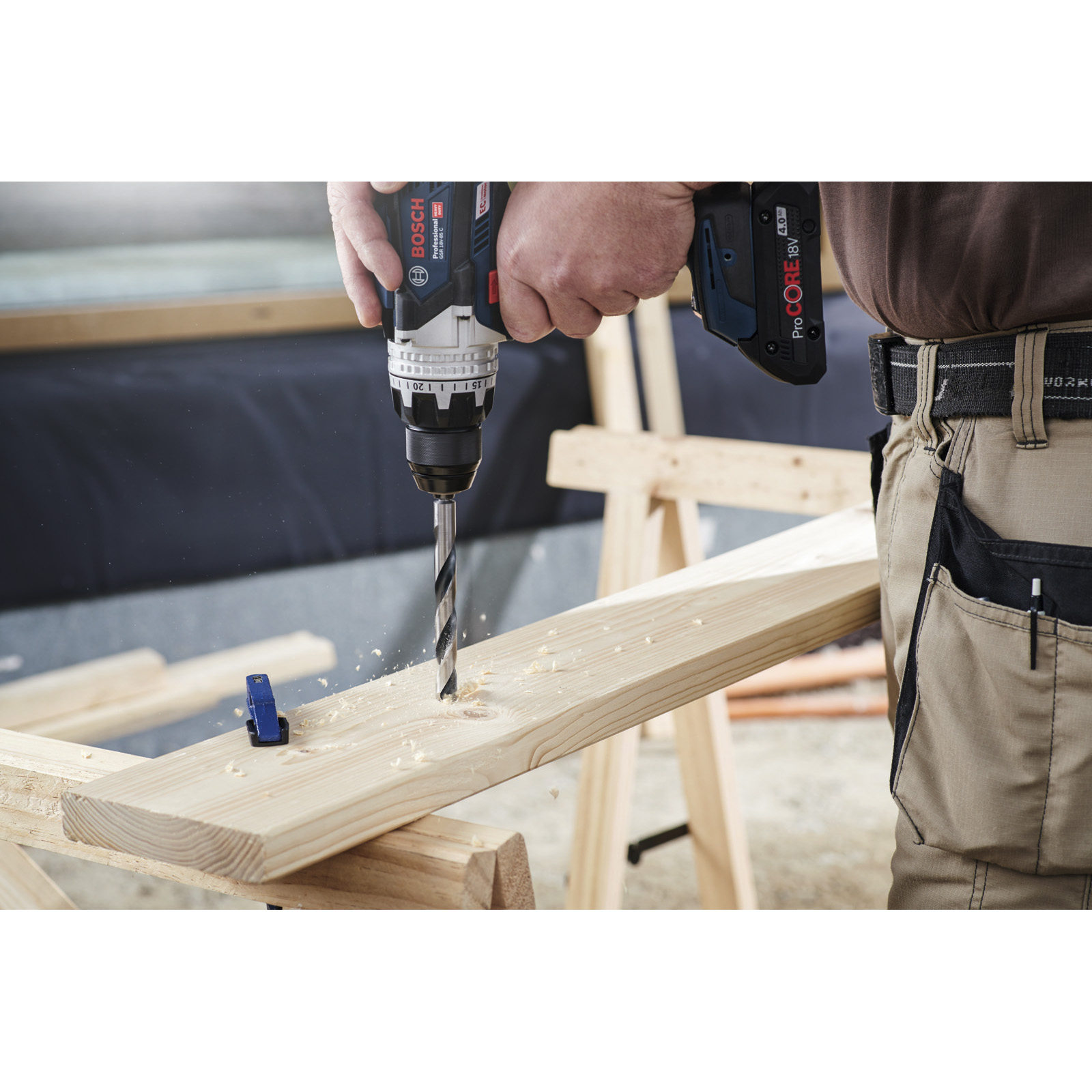 Osoba wierci drewnianą deskę za pomocą niebiesko-czarnej wiertarki. Widoczne są wióry drzewne. Tło jest rozmyte.