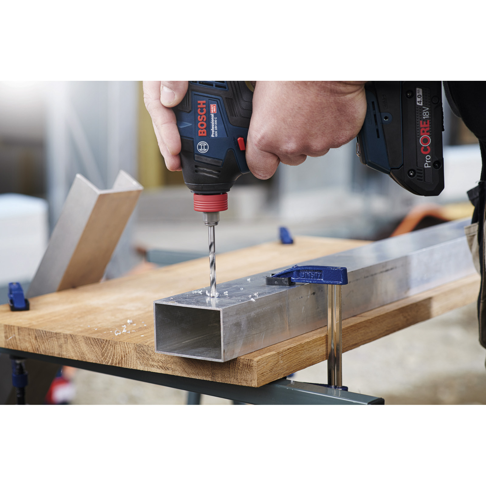 Osoba wierci metalową belkę za pomocą niebiesko-czarnej wiertarki. Drewno podpiera belkę. Tło jest rozmyte.