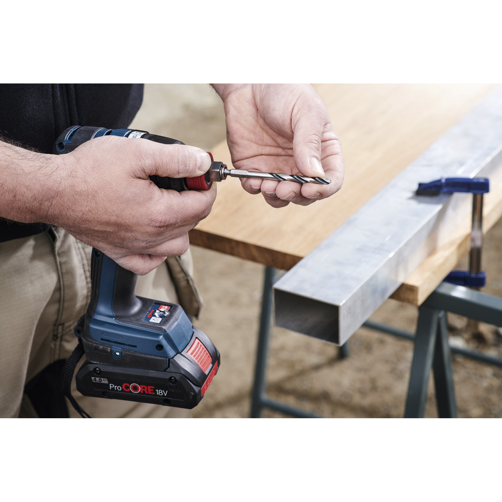 Osoba trzyma wiertarkę i wiertło, gotowa do wiercenia w prostokątnym metalowym pręcie.