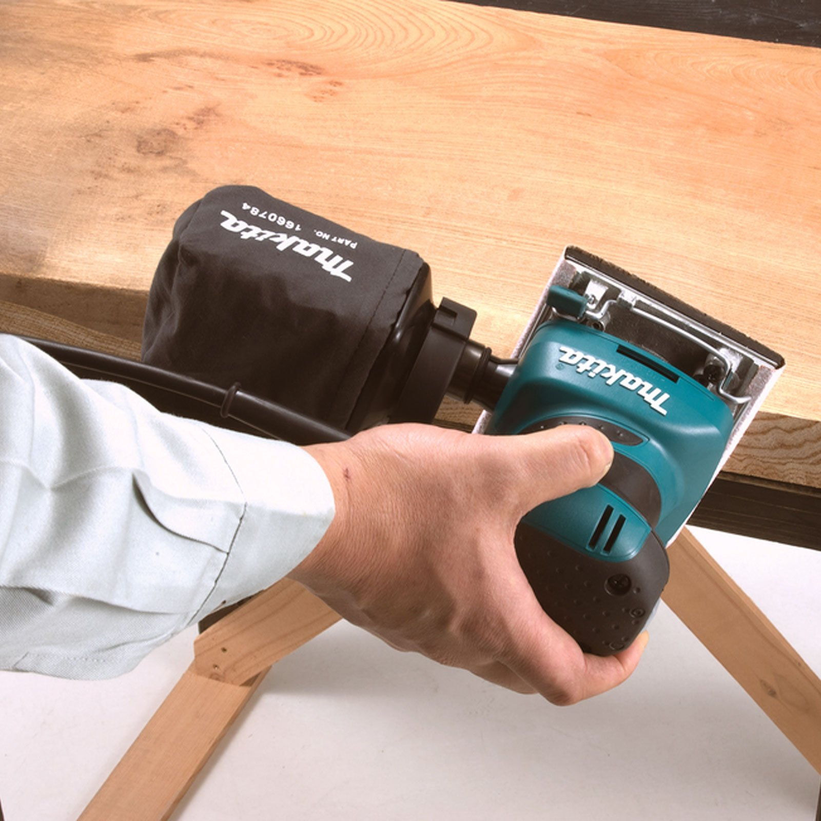 Una persona usa una levigatrice su una superficie di legno. La levigatrice è verde acqua e nera.