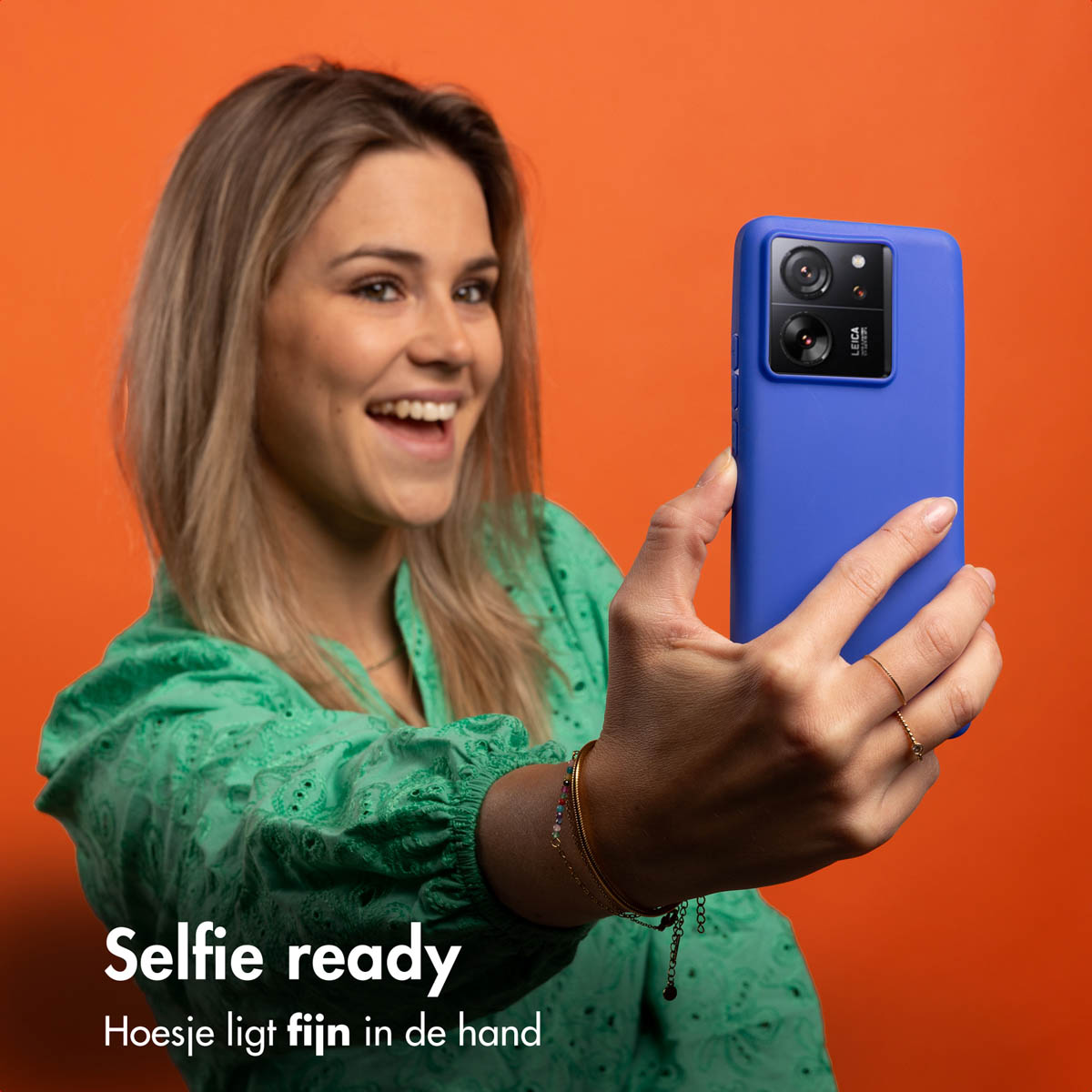 Frau macht Selfie mit Telefon in blauer Hülle. Sie hat blondes Haar und lächelt. Oranger Hintergrund.