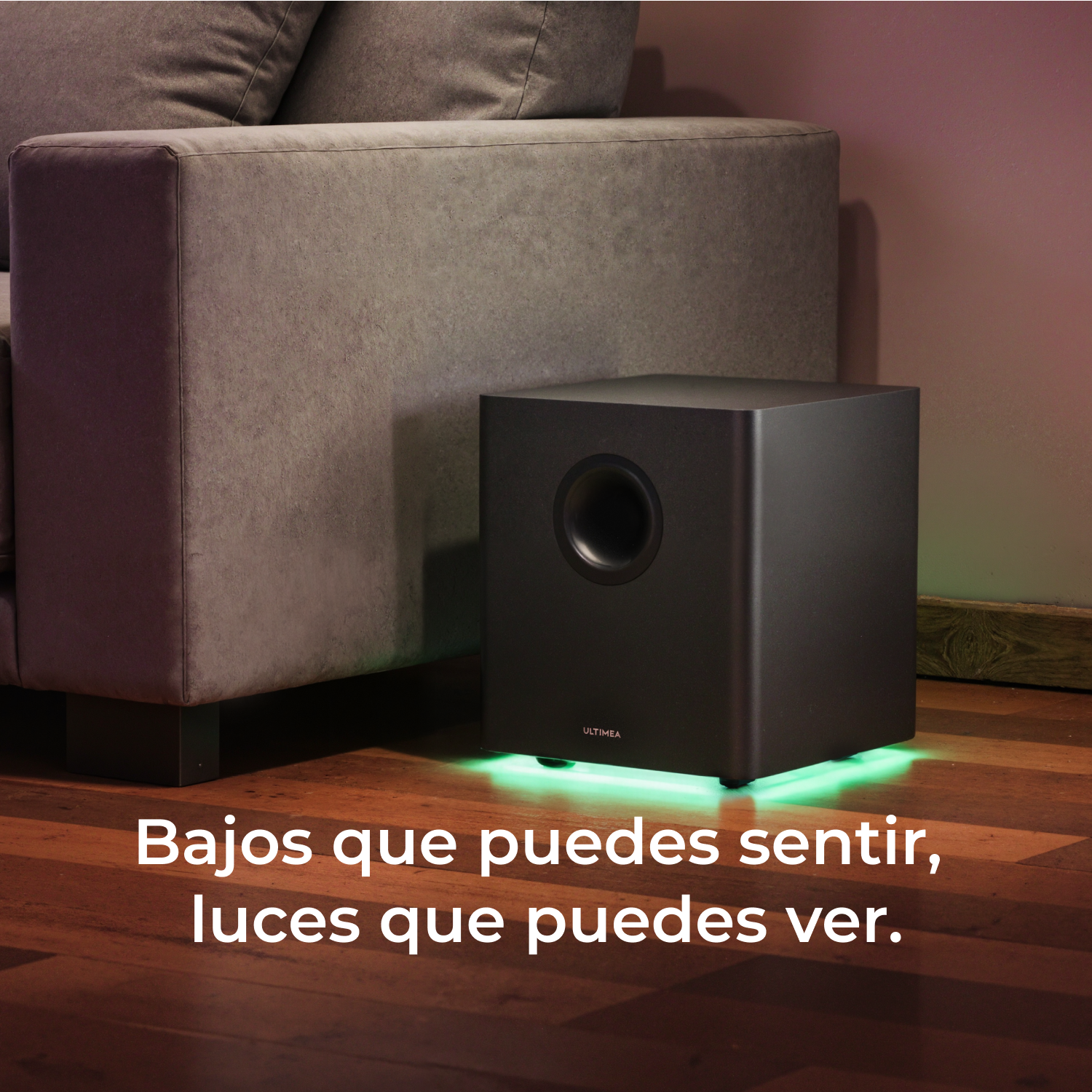 Un subwoofer negro junto a un sofá gris. Superposición de texto: Bajos que puedes sentir, luces que puedes ver.