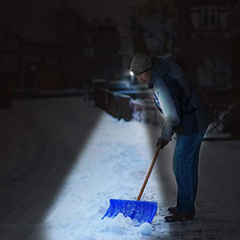 Ein Mann mit Hut und Licht schaufelt nachts Schnee. Das Licht des Hutes beleuchtet den Boden.