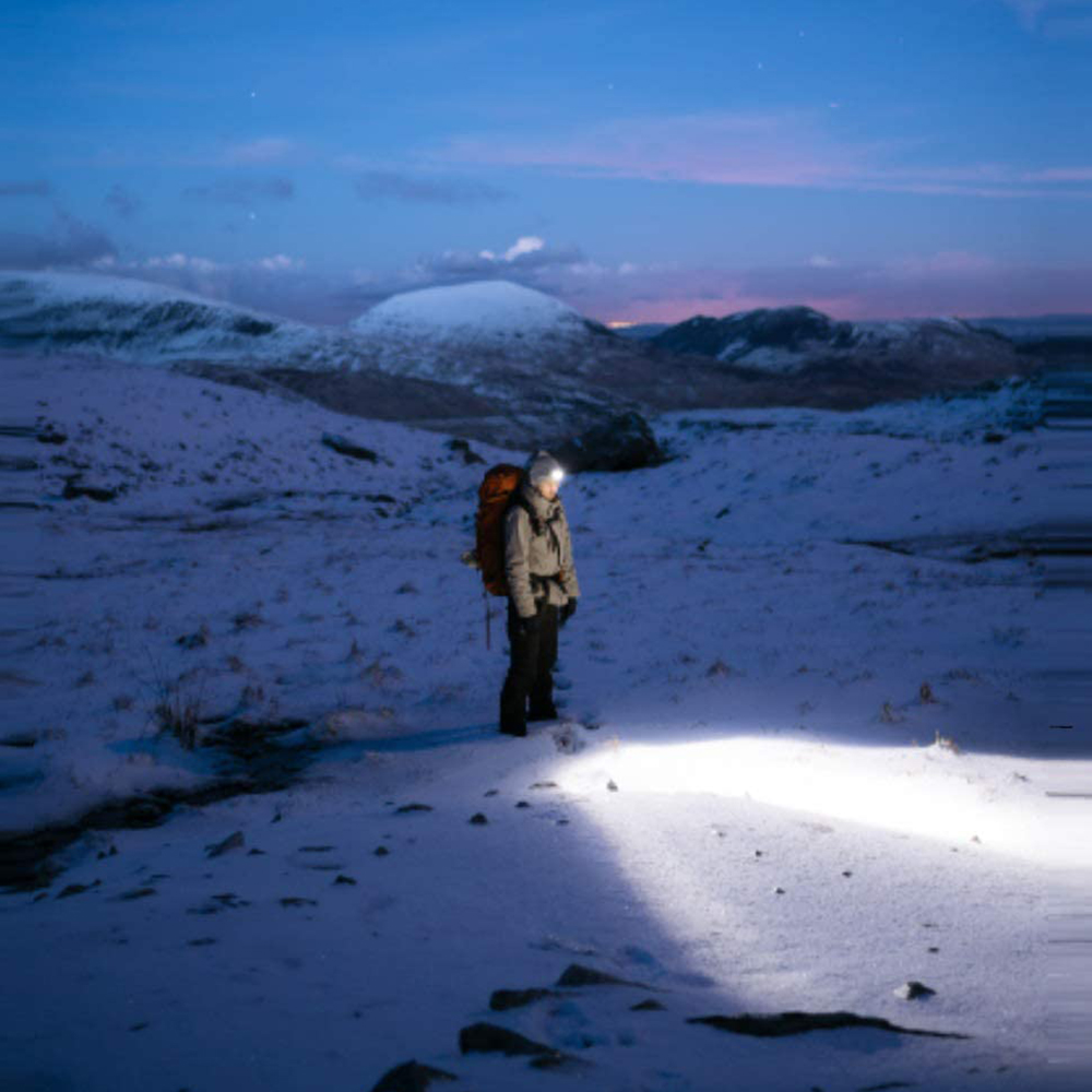 Person in schneebedeckter Landschaft mit Stirnlampe. Ferne Berge unter Dämmerungshimmel. Helles Licht beleuchtet den Vordergrund.