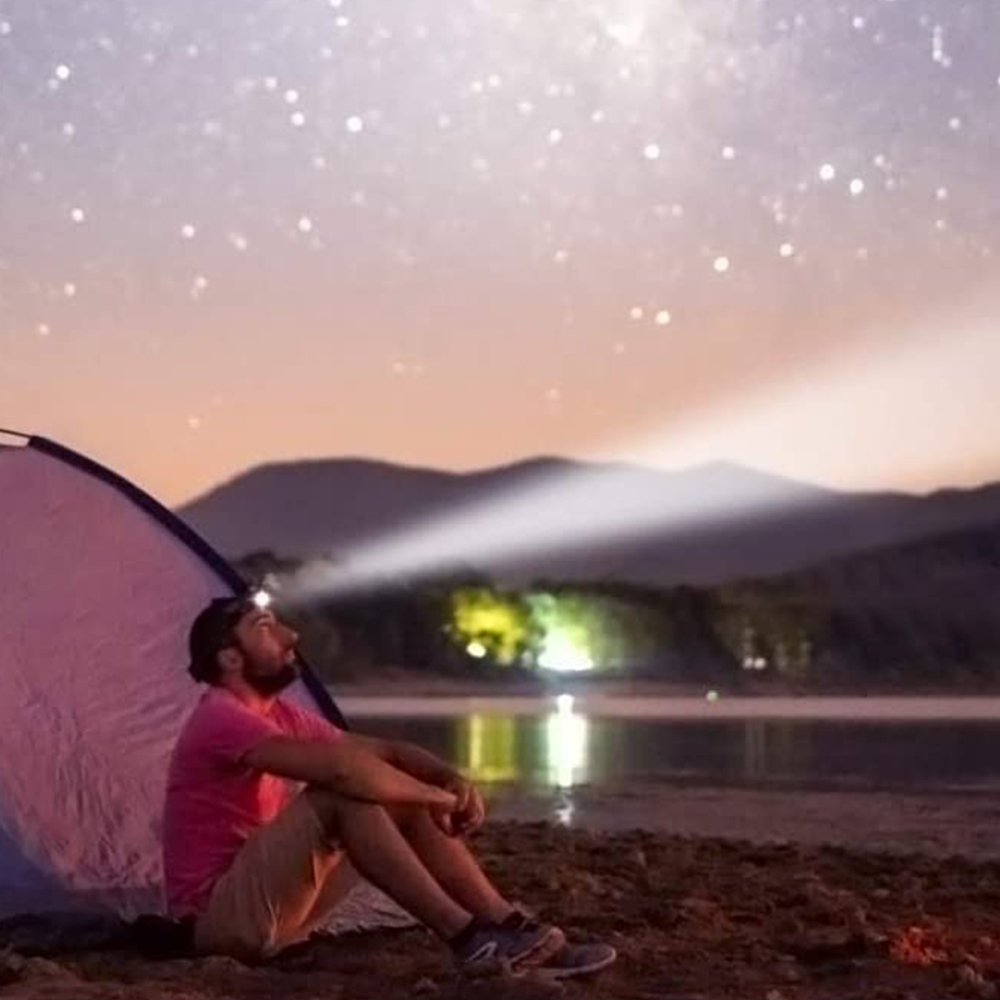 Person sitzt in der Nähe eines Zeltes und betrachtet den Sternenhimmel. Eine Stirnlampe beleuchtet die Szene.