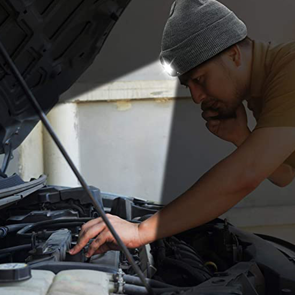 Ein Mann mit einer grauen Mütze mit Licht arbeitet an einem Automotor. Das Licht ist an.