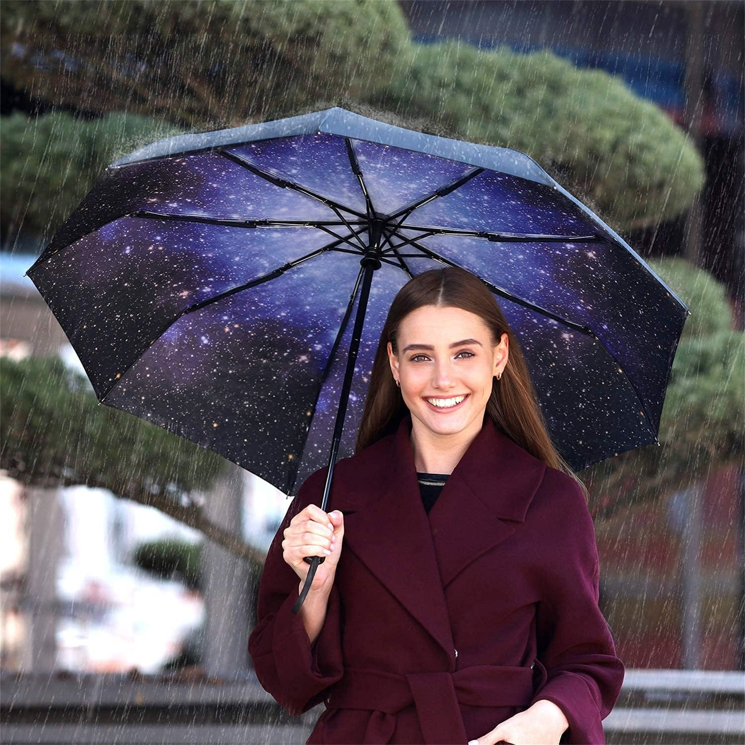 Eine Frau hält einen offenen Regenschirm mit Galaxien-Design im Regen und lächelt in die Kamera.