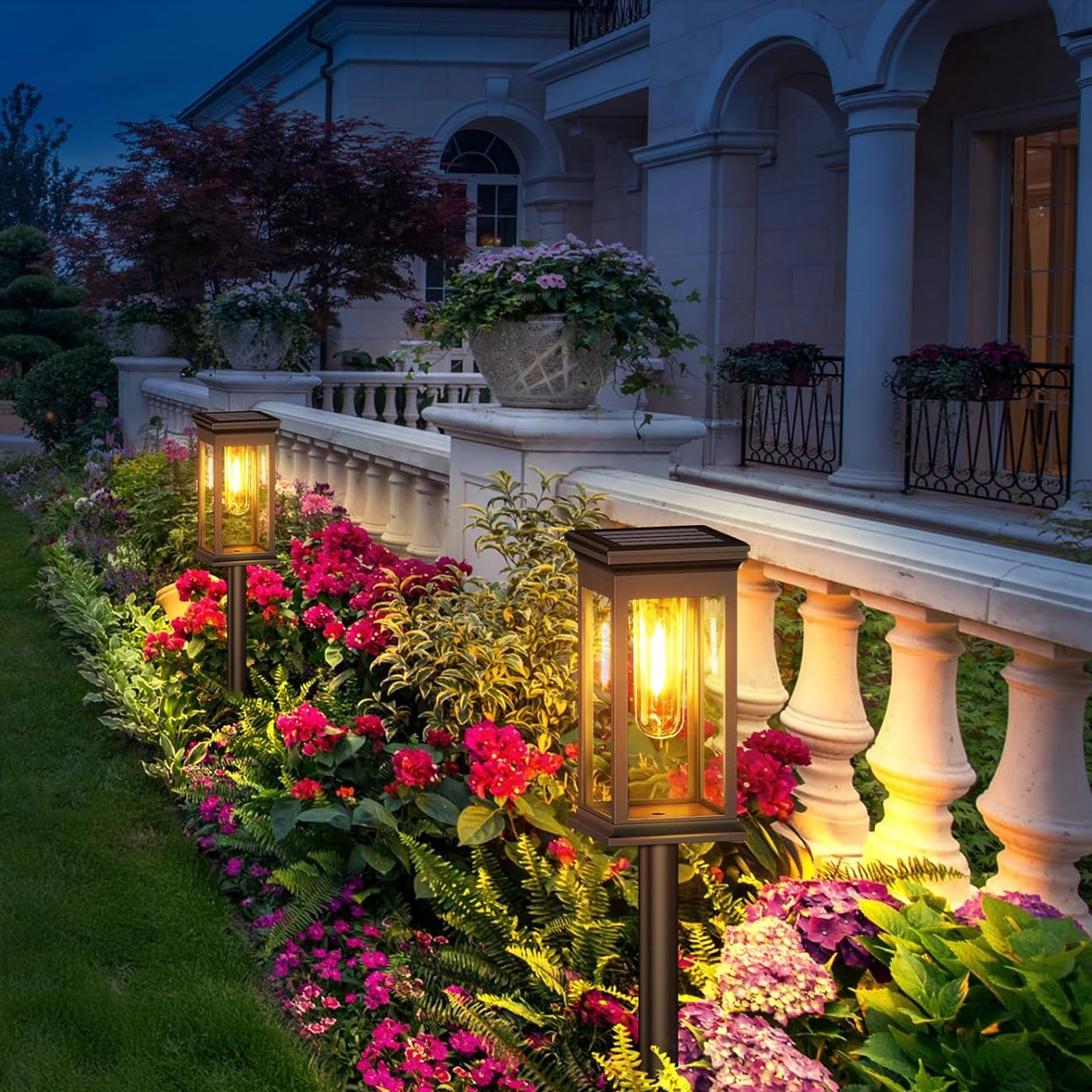 Zwei Solar-Gartenleuchten bei Nacht, die Blumen und ein weißes Gebäude beleuchten. Die Lichter sind schwarz und rechteckig.