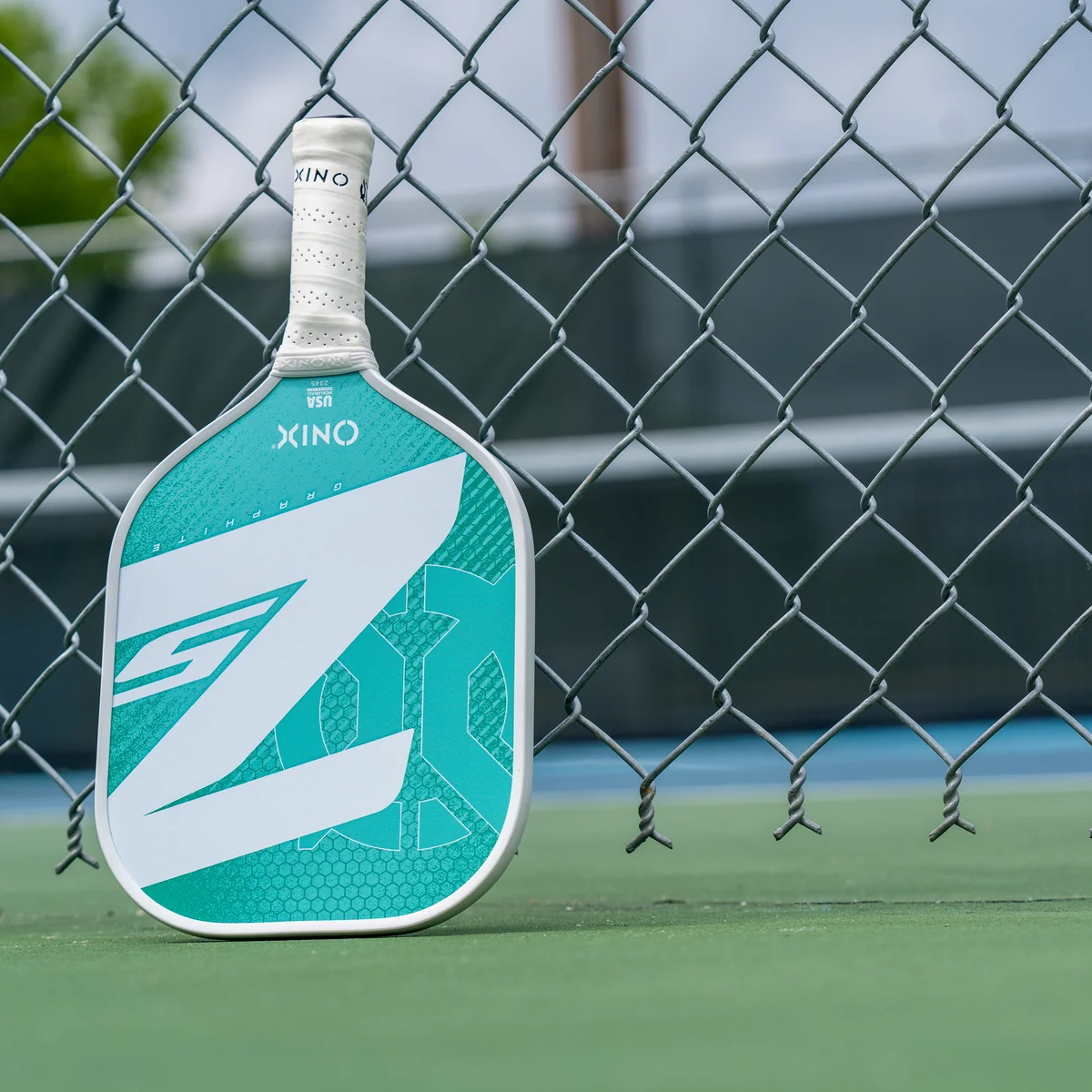 Ein weißer und blaugrüner Pickleball-Schläger lehnt an einem Maschendrahtzaun.