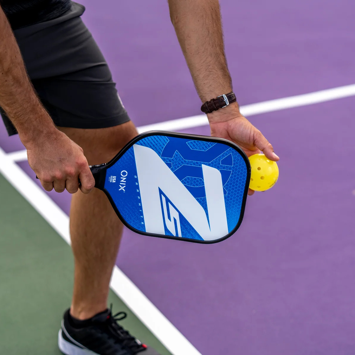 Eine Person hält einen blauen Pickleball-Schläger und einen gelben Ball auf einem lila Platz. Mann trägt Shorts.
