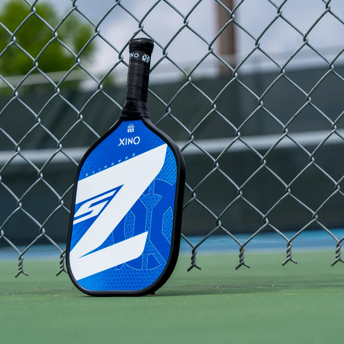 Ein Pickleball-Schläger steht an einem Zaun, wobei die Marke und das Logo auf dem Schläger sichtbar sind.