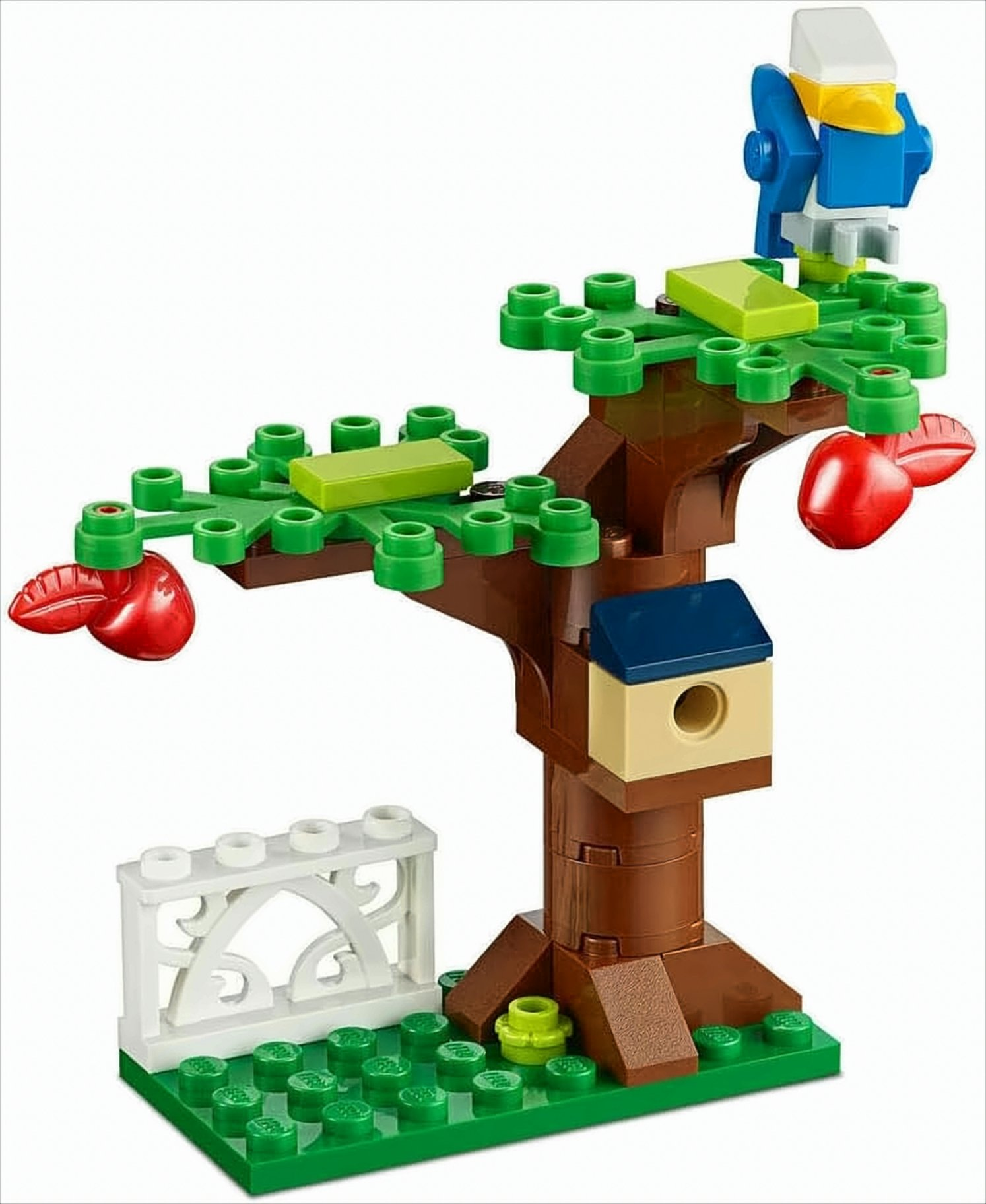 Ein Lego-Baum mit einem blauen Vogel, einem Vogelhaus und roten Äpfeln. Ein weißer Zaun im Vordergrund.