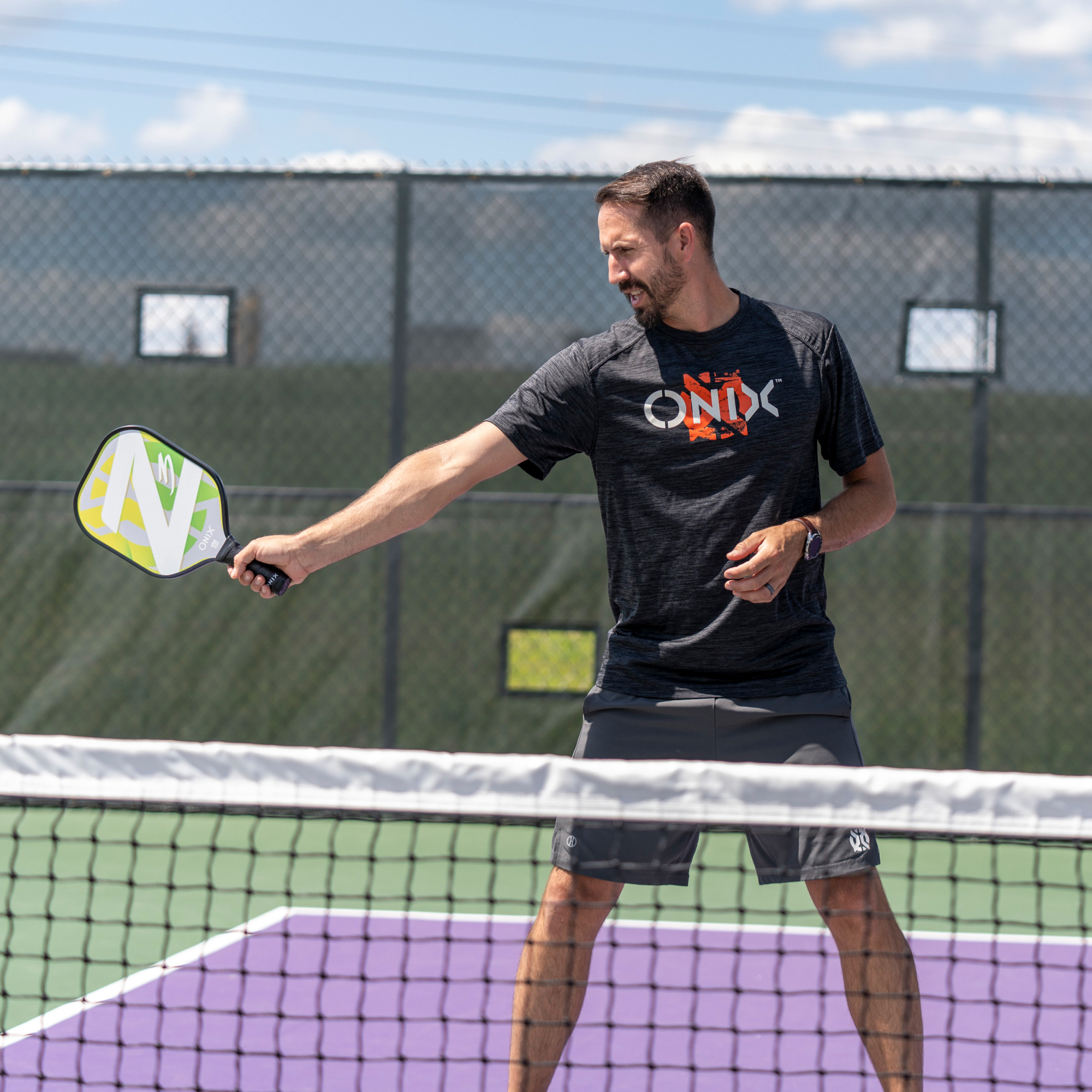Ein Mann spielt Pickleball, hält einen Schläger, weg von der Kamera. Grüner Platz und Netz sichtbar.