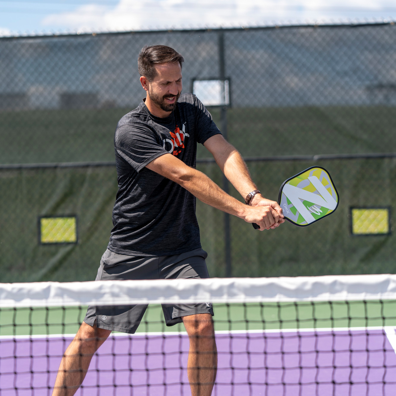 Ein Mann in einem schwarzen Hemd spielt Pickleball. Das Netz und der Platz sind im Vordergrund.