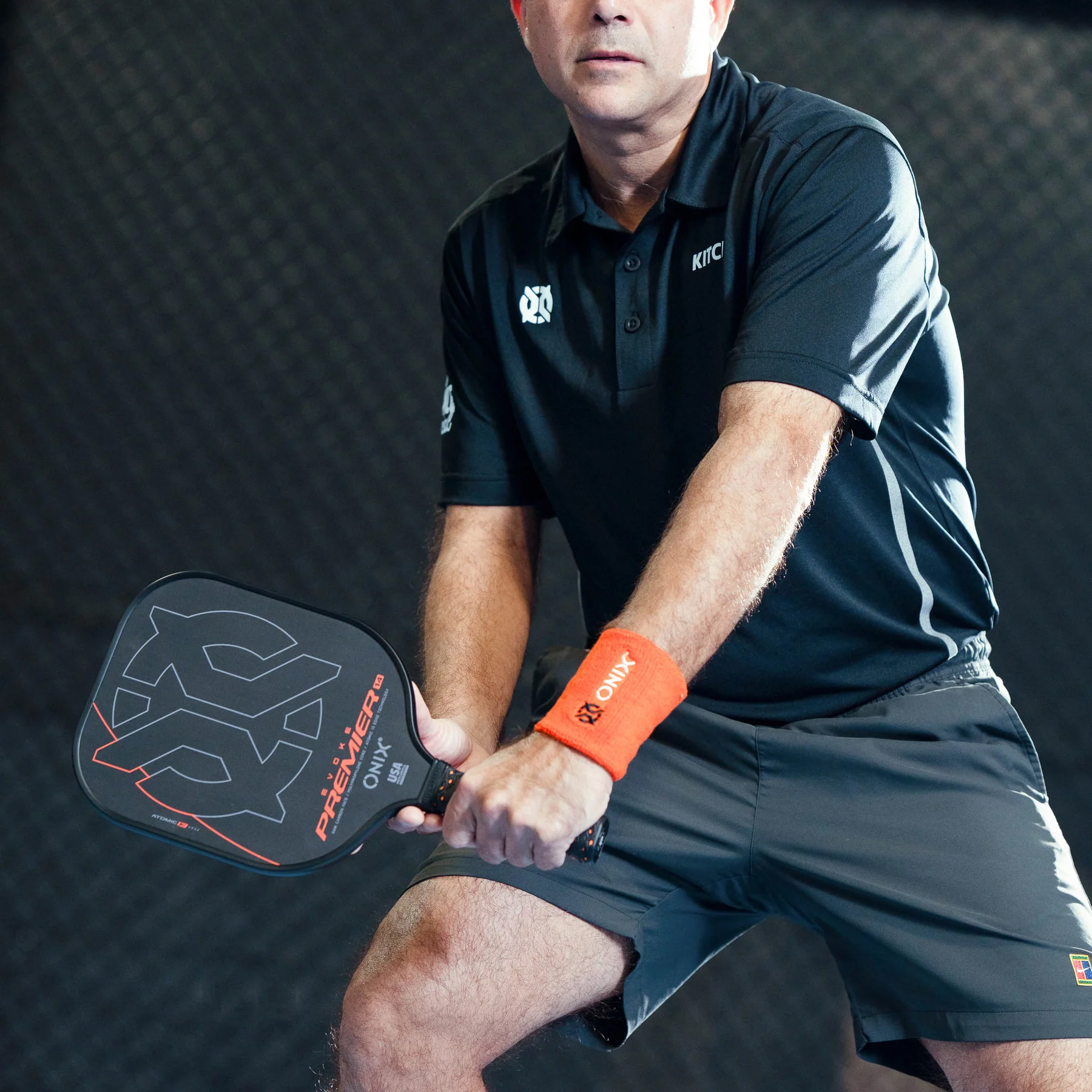Mann spielt Pickleball, hält einen Schläger. Er trägt ein schwarzes Hemd, graue Shorts und ein orangefarbenes Armband.