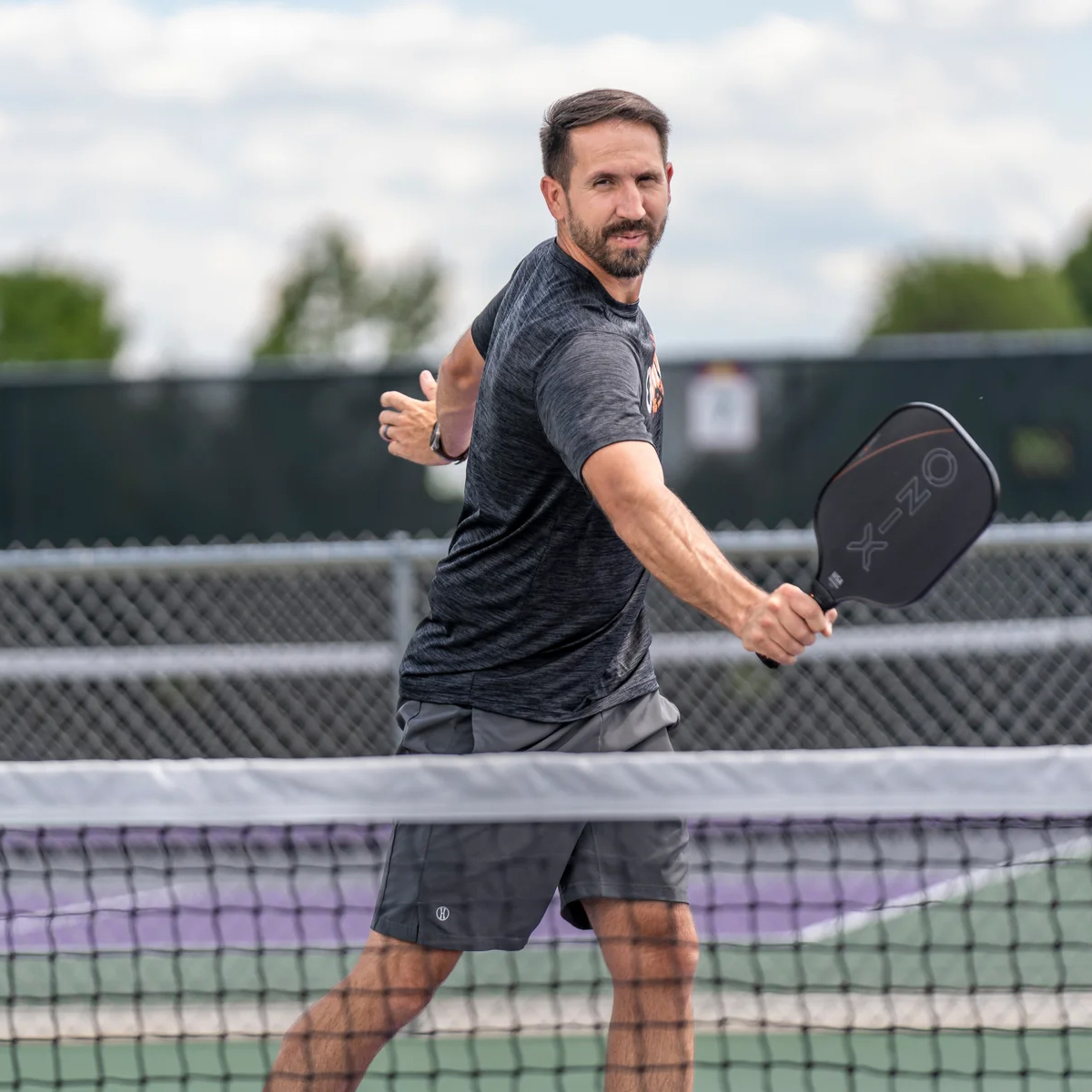 Ein Mann spielt Pickleball, hält einen schwarzen Schläger, in der Nähe eines Netzes.