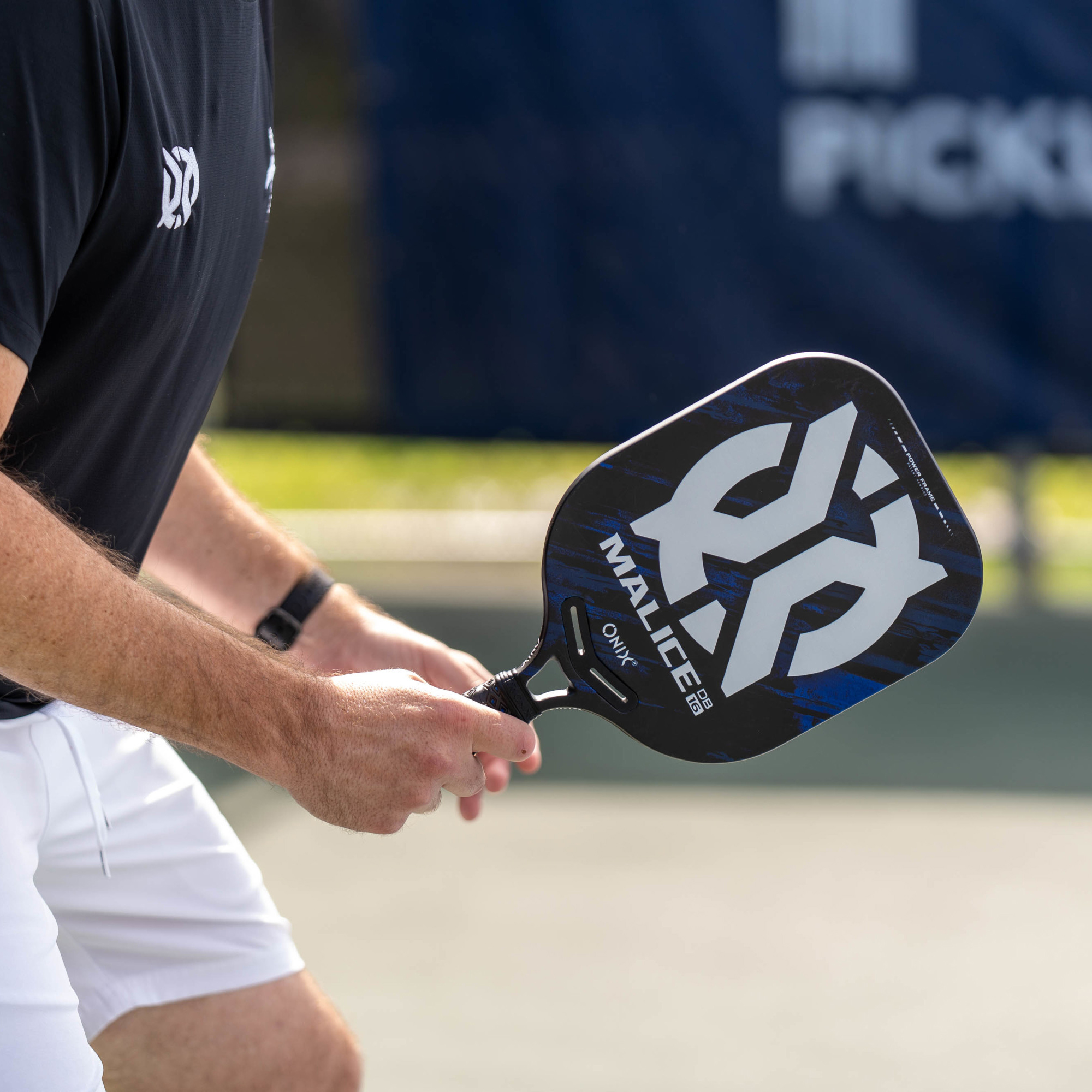 Eine Person hält einen Pickleball-Schläger mit schwarz-blauem Design. Der Hintergrund ist ein Pickleball-Feld.