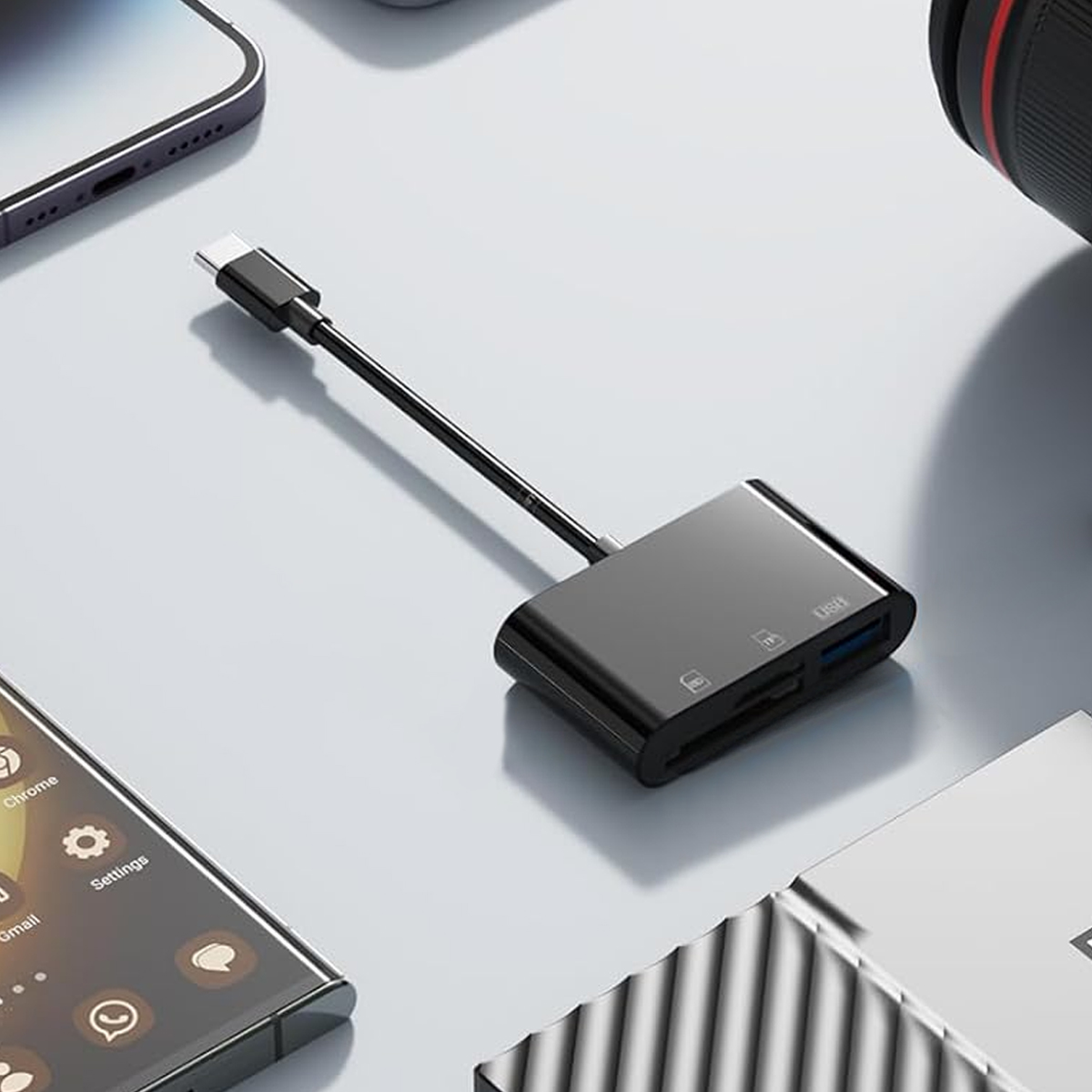 Adattatore USB-C nero con cavo nero, vicino a un telefono e a un obiettivo della fotocamera. Superficie grigia.