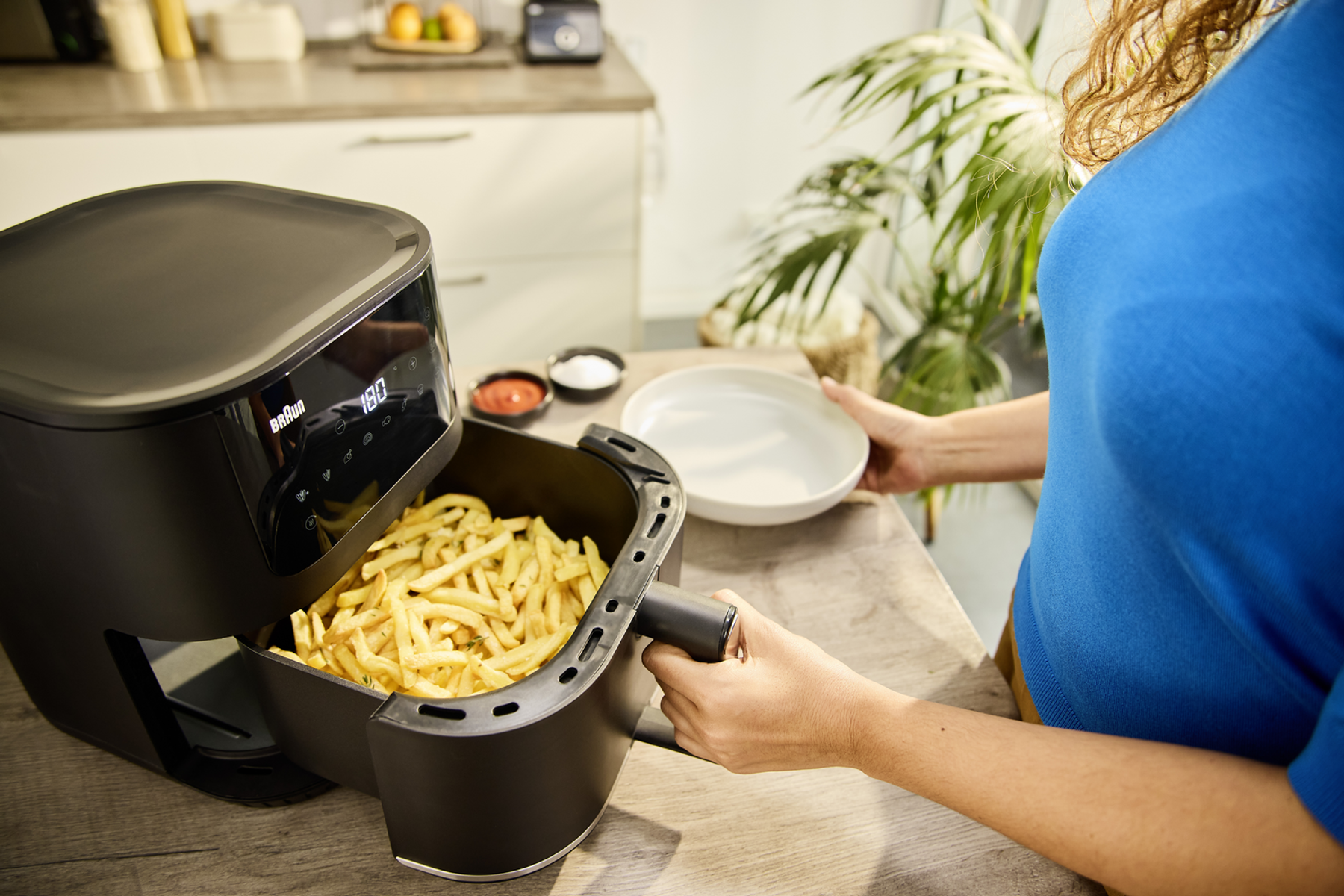 Kobieta wyjmuje frytki z czarnej frytownicy z białym talerzem. W pobliżu są sosy.