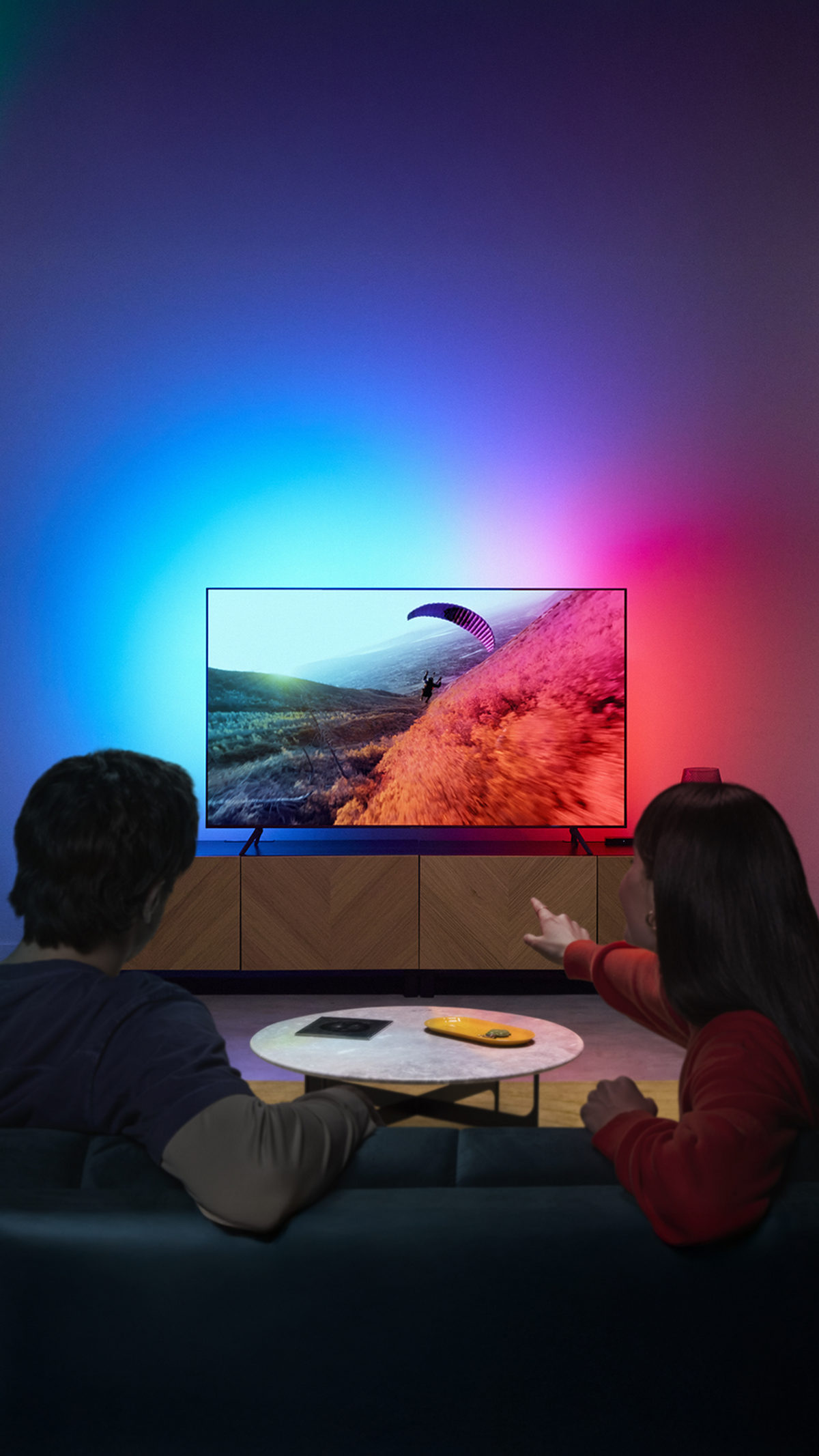 Çift, bir oturma odasında ortam ışıklarıyla TV'de yamaç paraşütçüsünü izliyor.