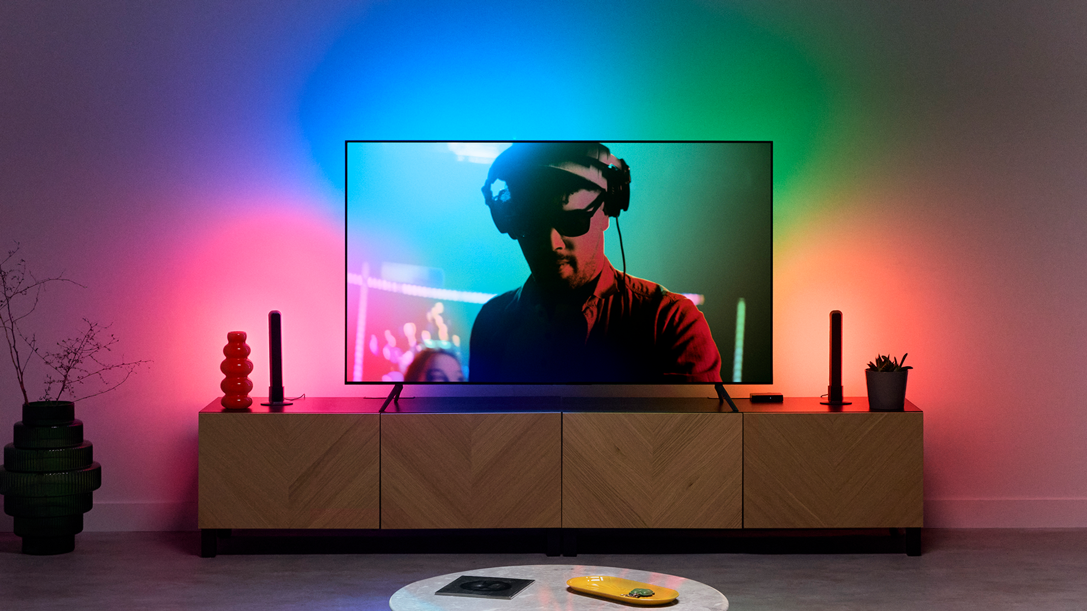 Kulaklıklı bir adam, ahşap bir dolapta renkli ışıklarla bir TV izliyor. Yerde sarı bir nesne var.