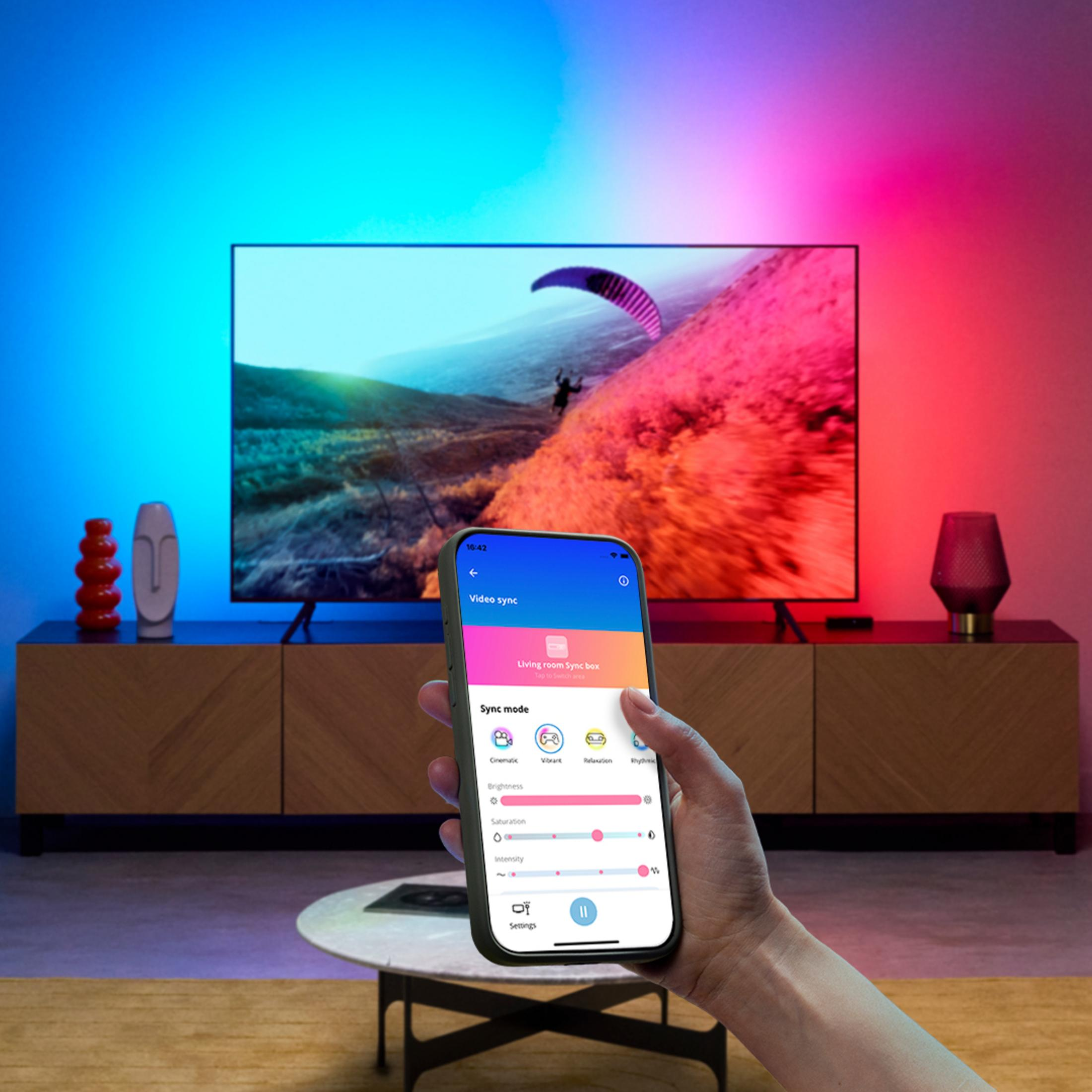 Bir kişinin eli, arka planda TV ve ışıklarla, bir senkronizasyon uygulaması gösteren bir akıllı telefon tutuyor.