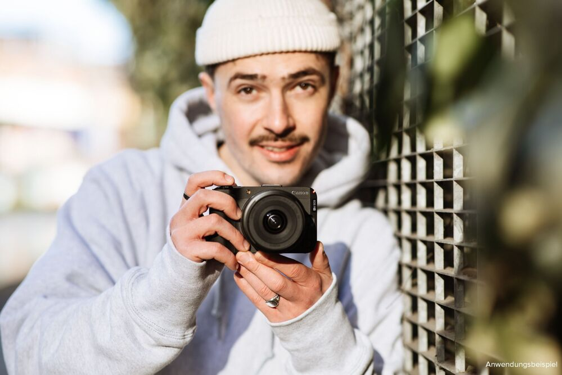 Mężczyzna trzymający czarny aparat, uśmiechający się do kamery. Ma na sobie białą czapkę i szarą bluzę z kapturem. Tło jest rozmyte.