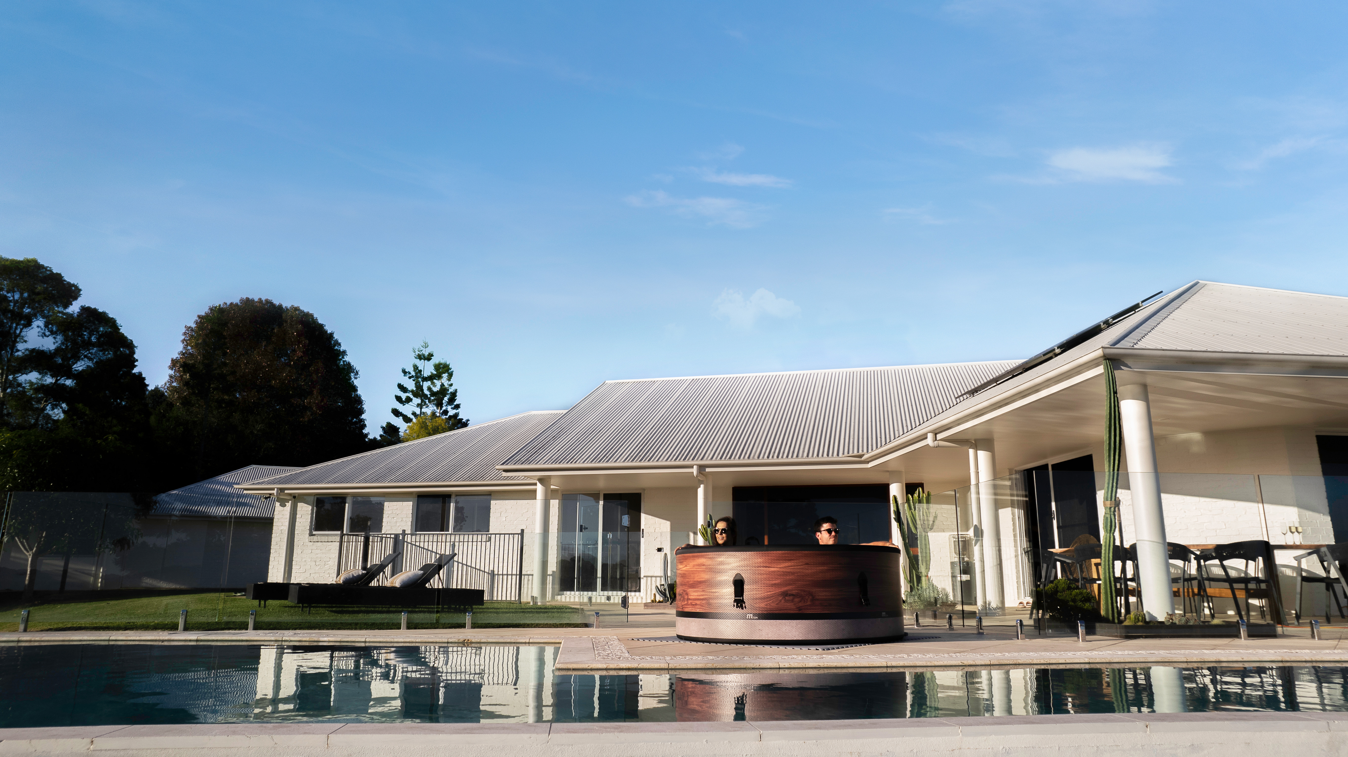 Außenaufnahme eines Hauses mit Pool. Zwei Personen sind in einem hölzernen Whirlpool. Blauer Himmel.
