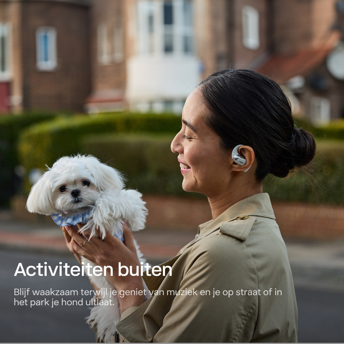 Kobieta trzyma małego białego psa, nosi słuchawki, uśmiecha się. Tło: ulica z domami. Tekst: Zajęcia na świeżym powietrzu.
