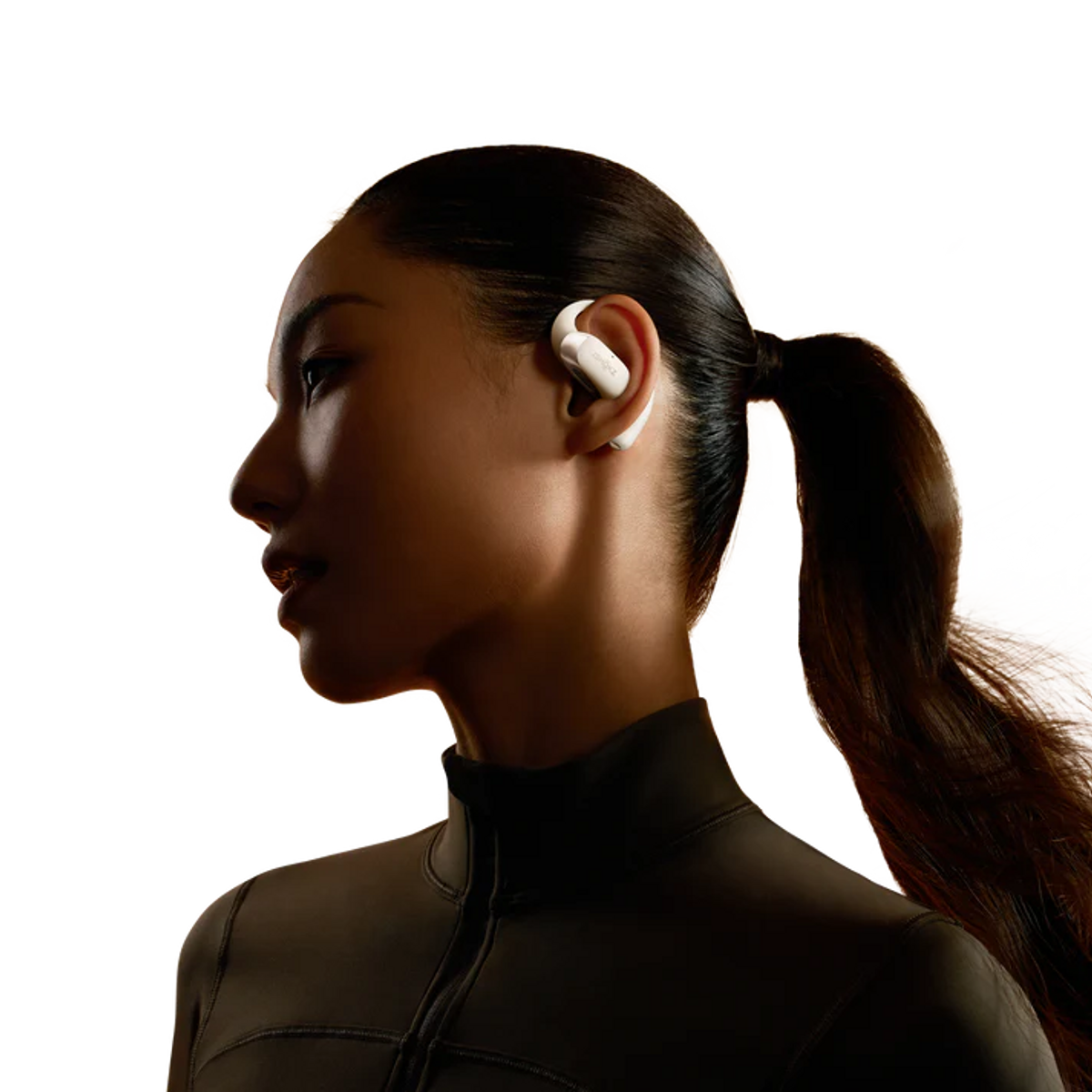 Mujer de perfil con un auricular blanco, fondo negro. Viste una parte superior oscura, mirando a la izquierda.