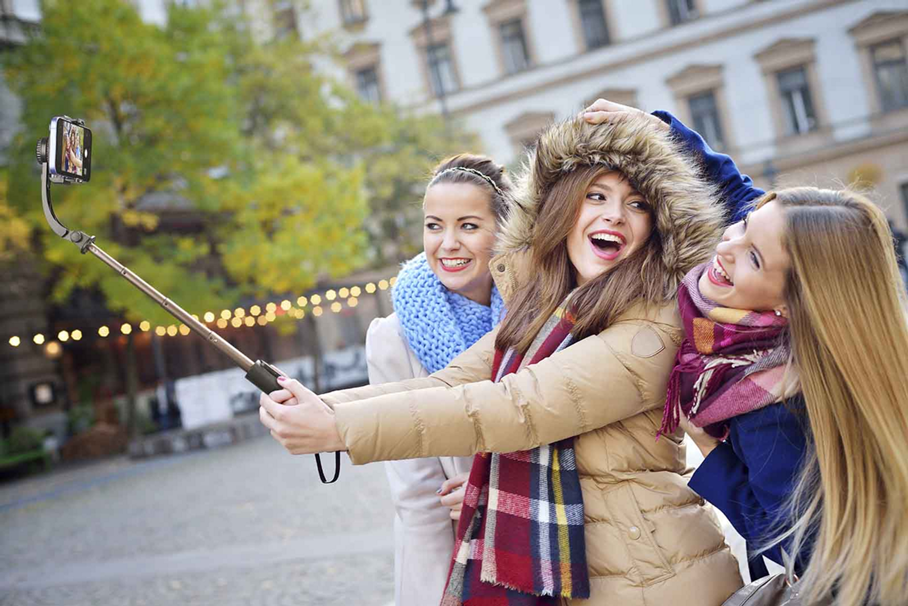 Drei Frauen machen ein Selfie mit einem Selfie-Stick und lachen. Hintergrund: Gebäude und Bäume.