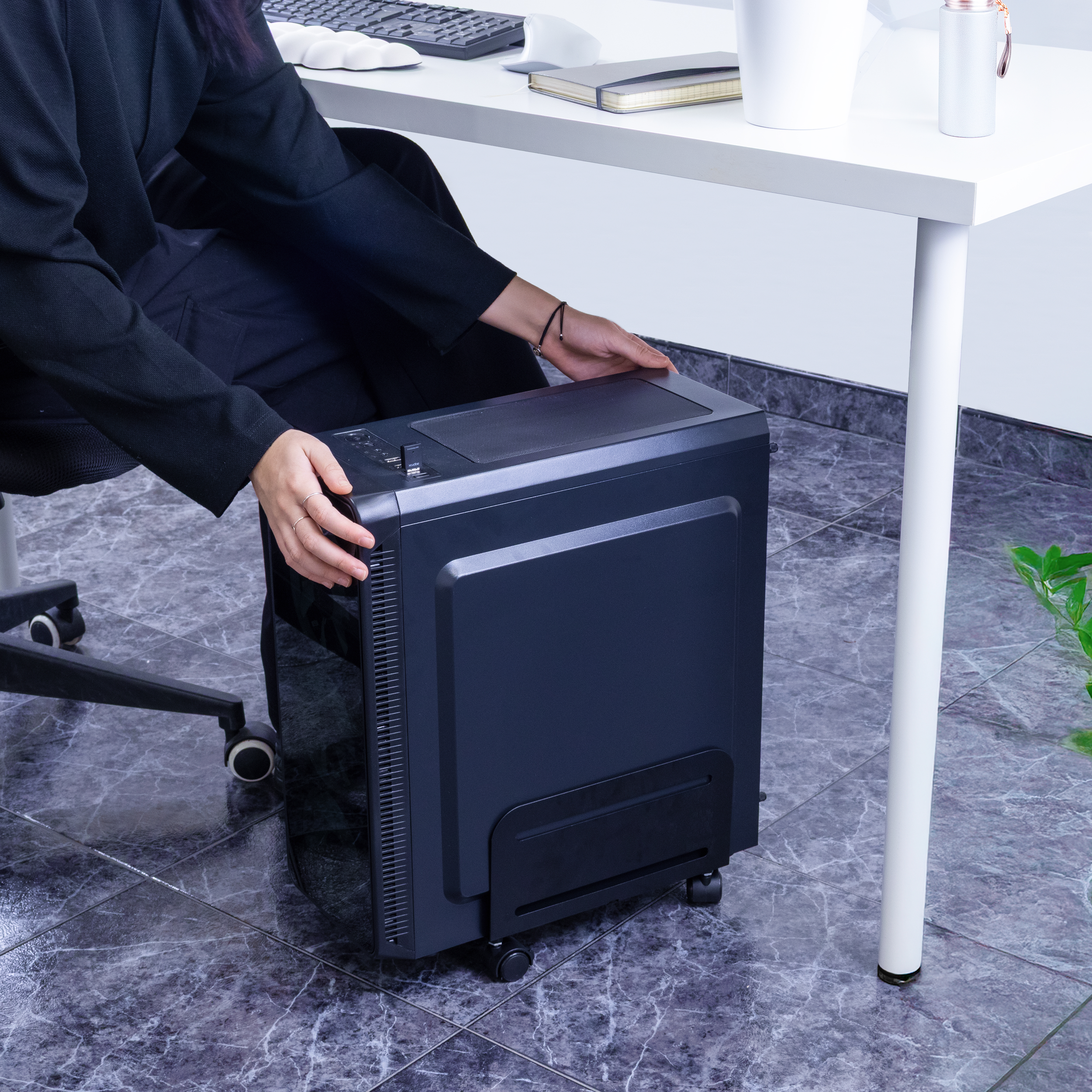 Persona colocando una torre de computadora en un soporte con ruedas debajo de un escritorio. El soporte es negro.
