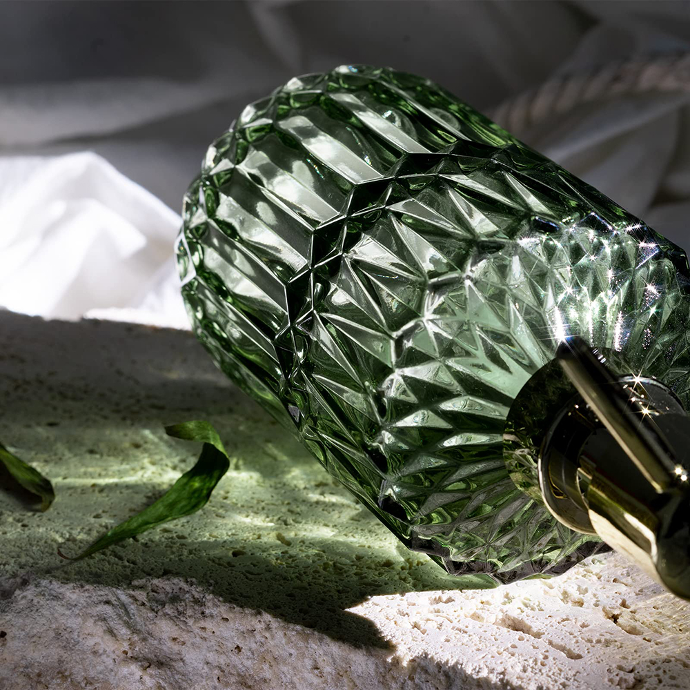 Eine grüne Glasflasche mit goldener Pumpe und strukturierter Oberfläche, die auf einer Steinoberfläche ruht.