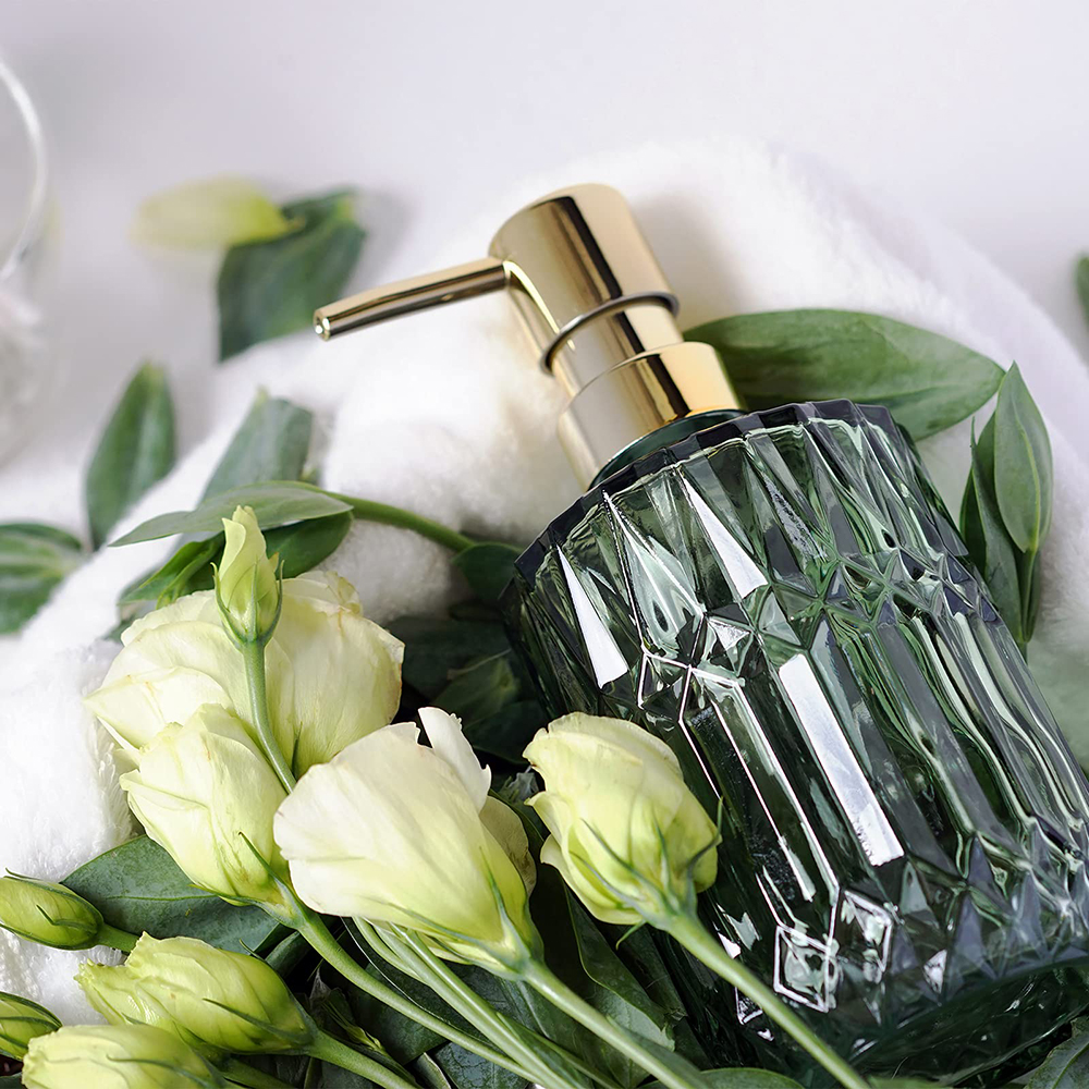 Grüner Glas-Seifenspender mit goldener Pumpe, weiße Blumen und grüne Blätter auf weißem Handtuch.