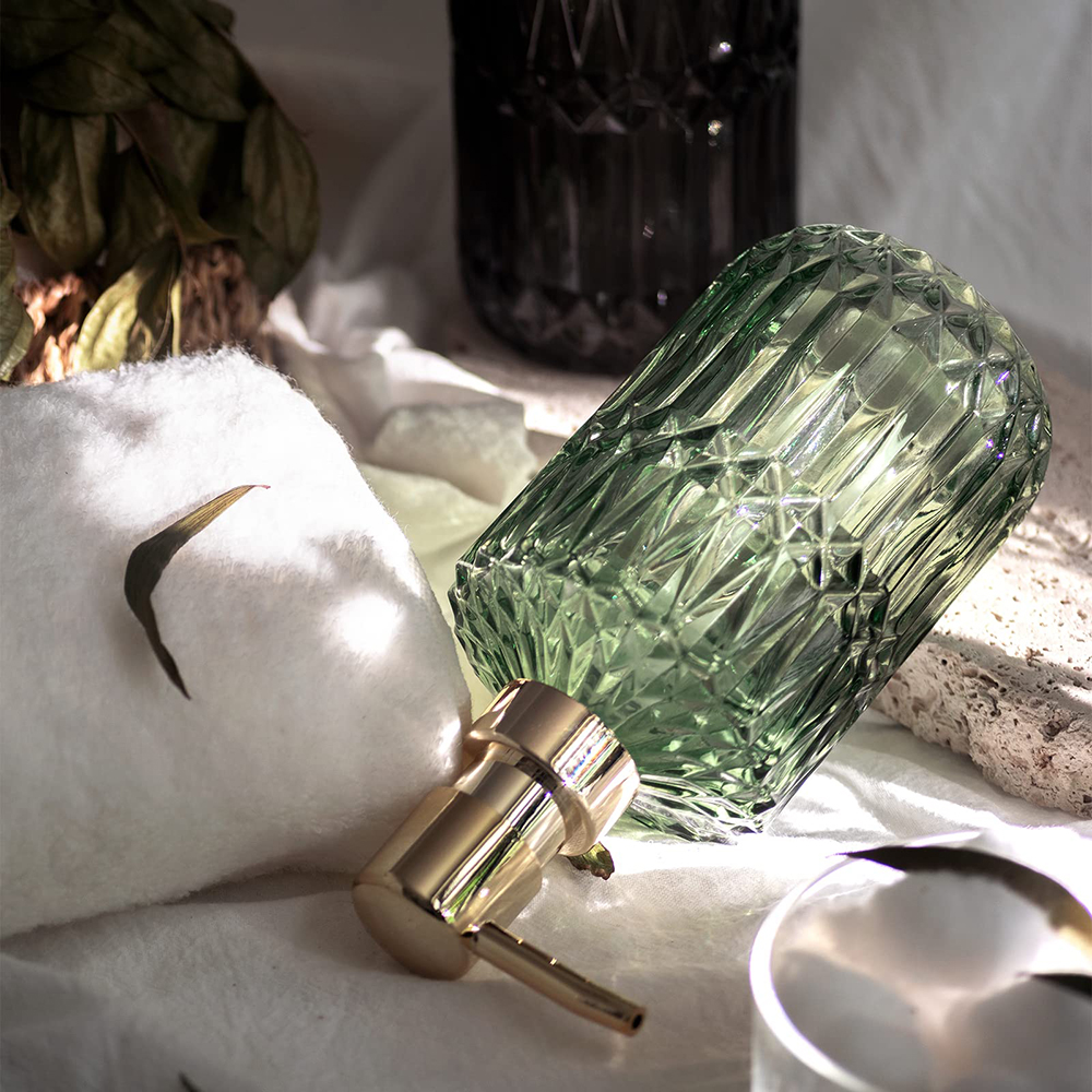 Eine grüne Glasflasche mit goldener Pumpe, neben einem weißen Handtuch und einer dunklen Glasflasche, auf einer weißen Oberfläche.