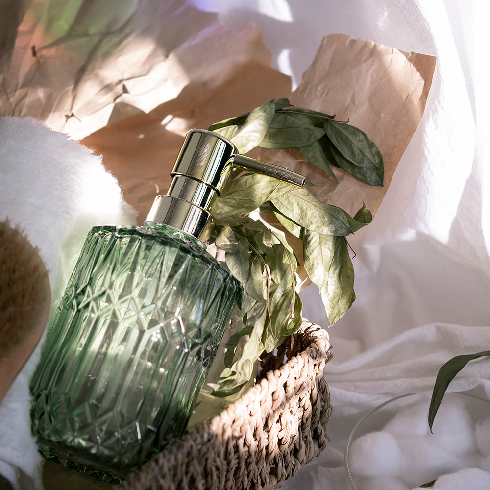 Grüner Glas-Seifenspender mit goldener Pumpe, Blättern und Korb auf weißer Oberfläche.