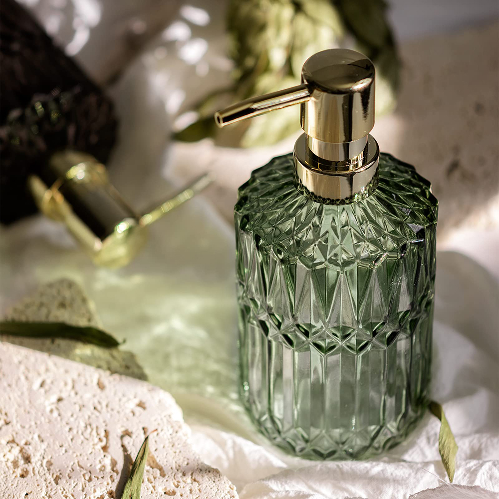 Grüner Glas-Seifenspender mit goldener Pumpe auf weißer Oberfläche, mit Steinen und Grünzeug drumherum.