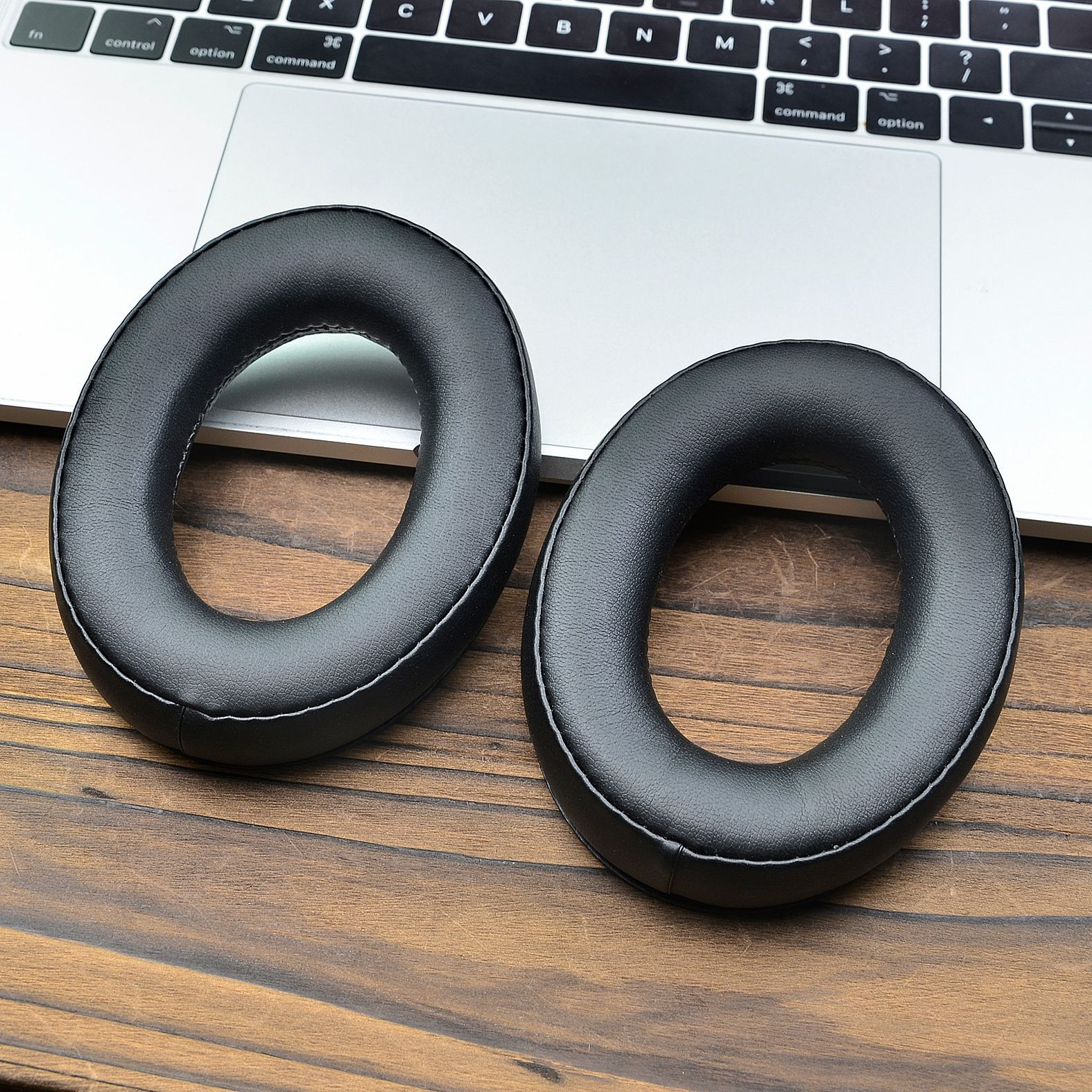 Due cuscinetti auricolari neri e rotondi per cuffie si trovano su una superficie di legno accanto alla tastiera di un laptop.