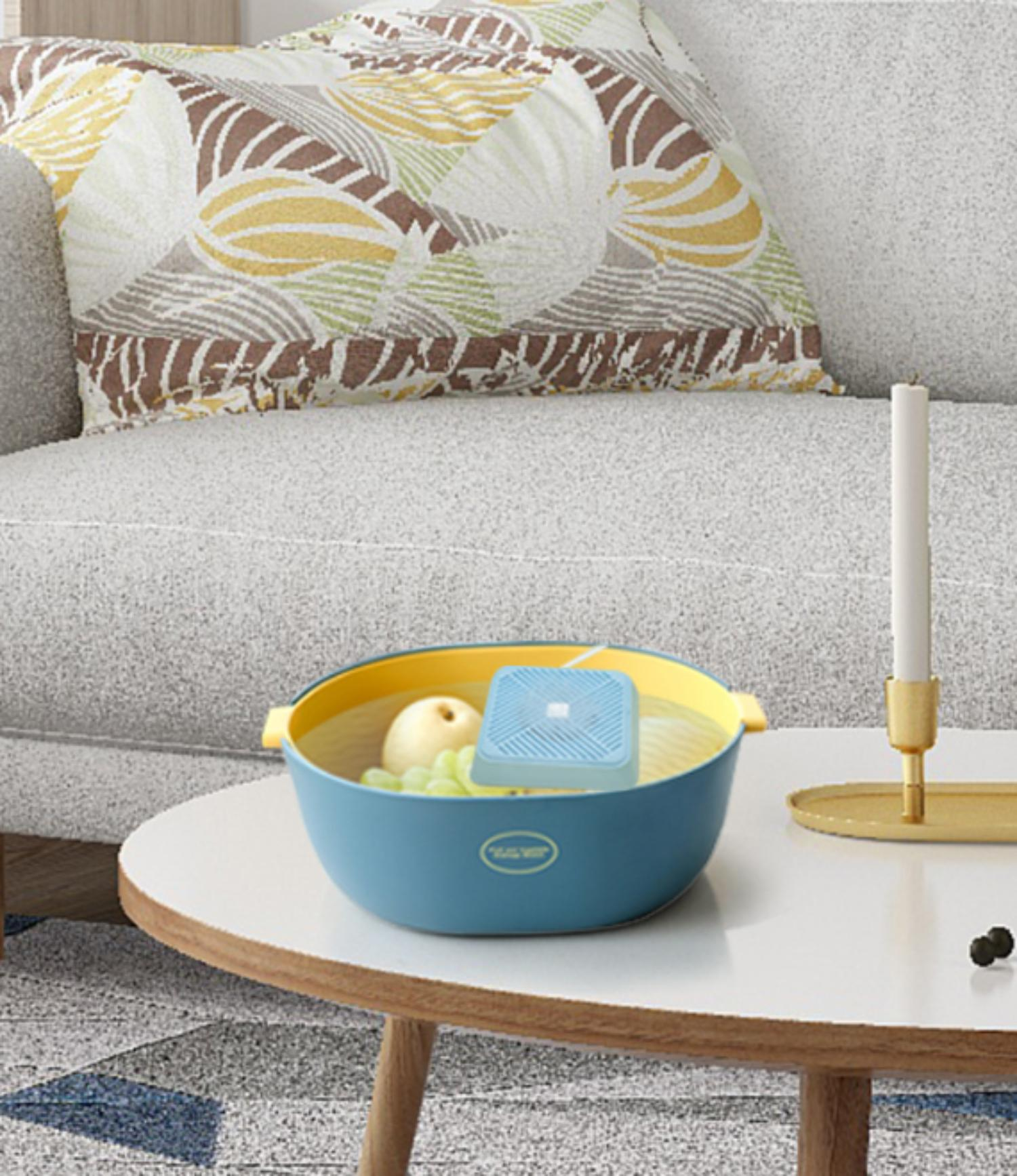 Eine blaue Schüssel mit einem Apfel und einem Gerät auf einem weißen Tisch, mit einem Sofa im Hintergrund.
