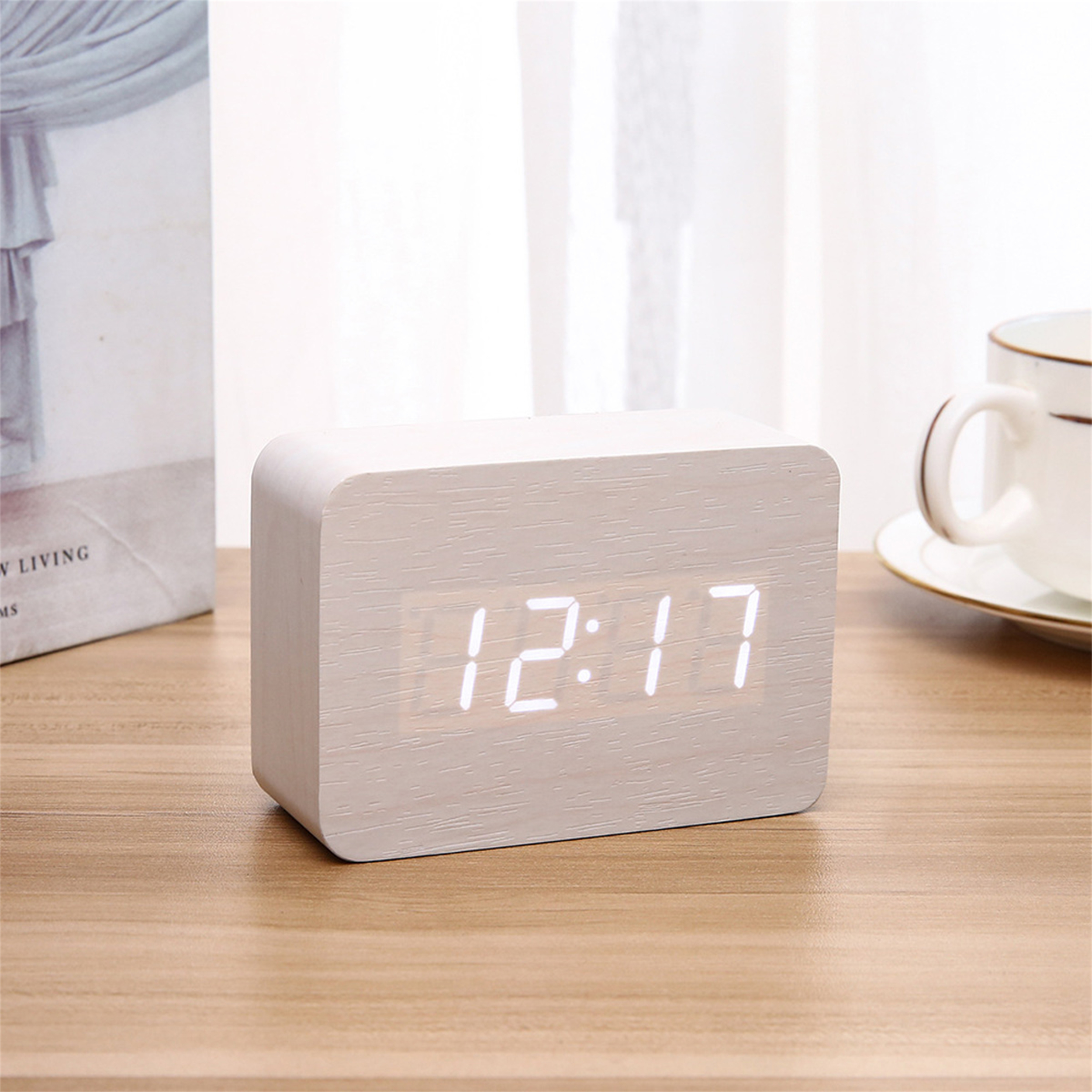 Eine Holzuhr zeigt "12:17" an. Sie steht auf einem Holztisch, im Hintergrund ein Buch und eine Tasse.