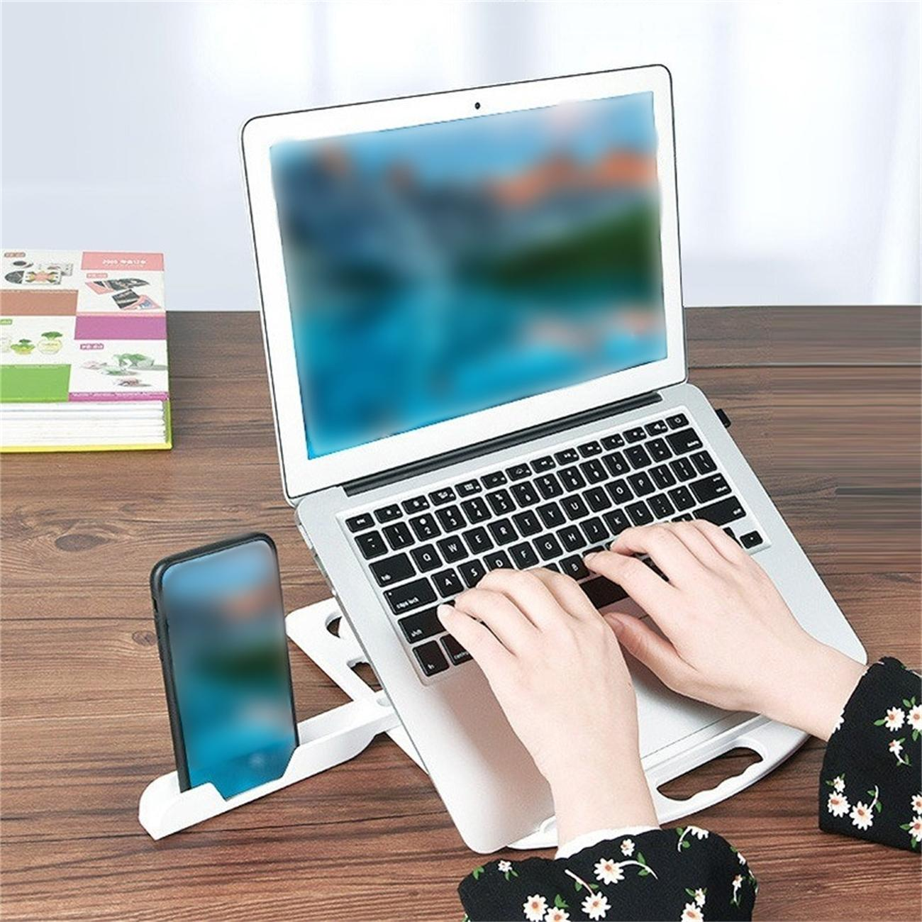 Hände tippen auf Laptop auf einem weißen Laptopständer mit einem Telefon. Bücher sind im Hintergrund.