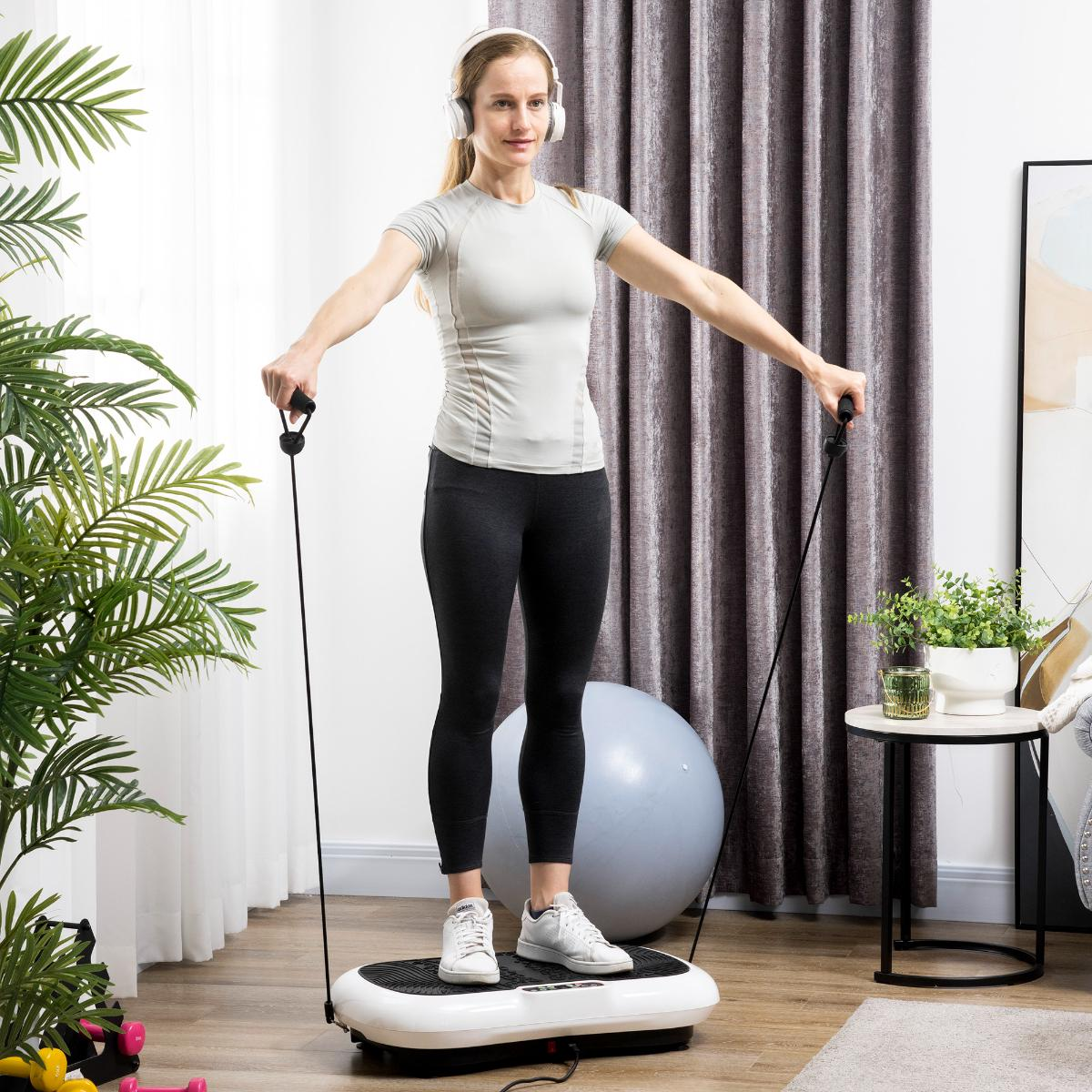 Frau mit Kopfhörern steht auf einer Vibrationsplatte und hält Trainingsseile.