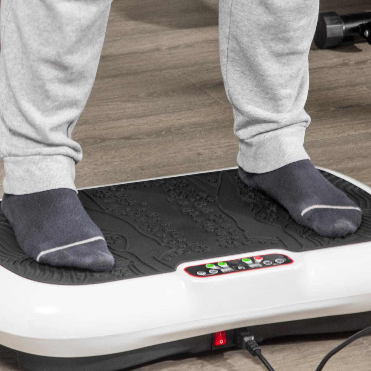 Füße einer Person auf einer weißen und schwarzen Vibrationsplattform. Person trägt graue Jogginghose und dunkle Socken.