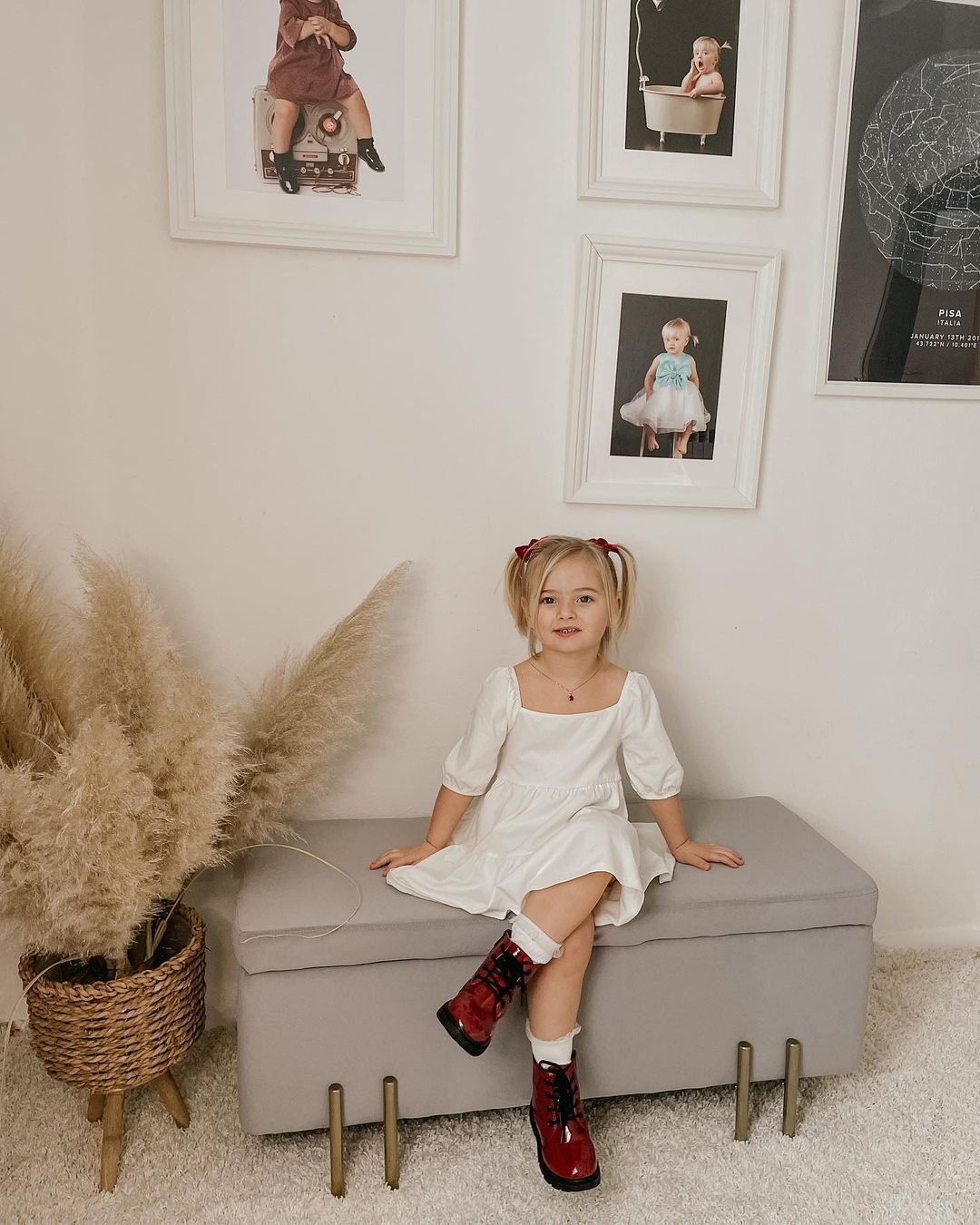 Una bambina in abito bianco seduta su una panca grigia, foto incorniciate al muro.