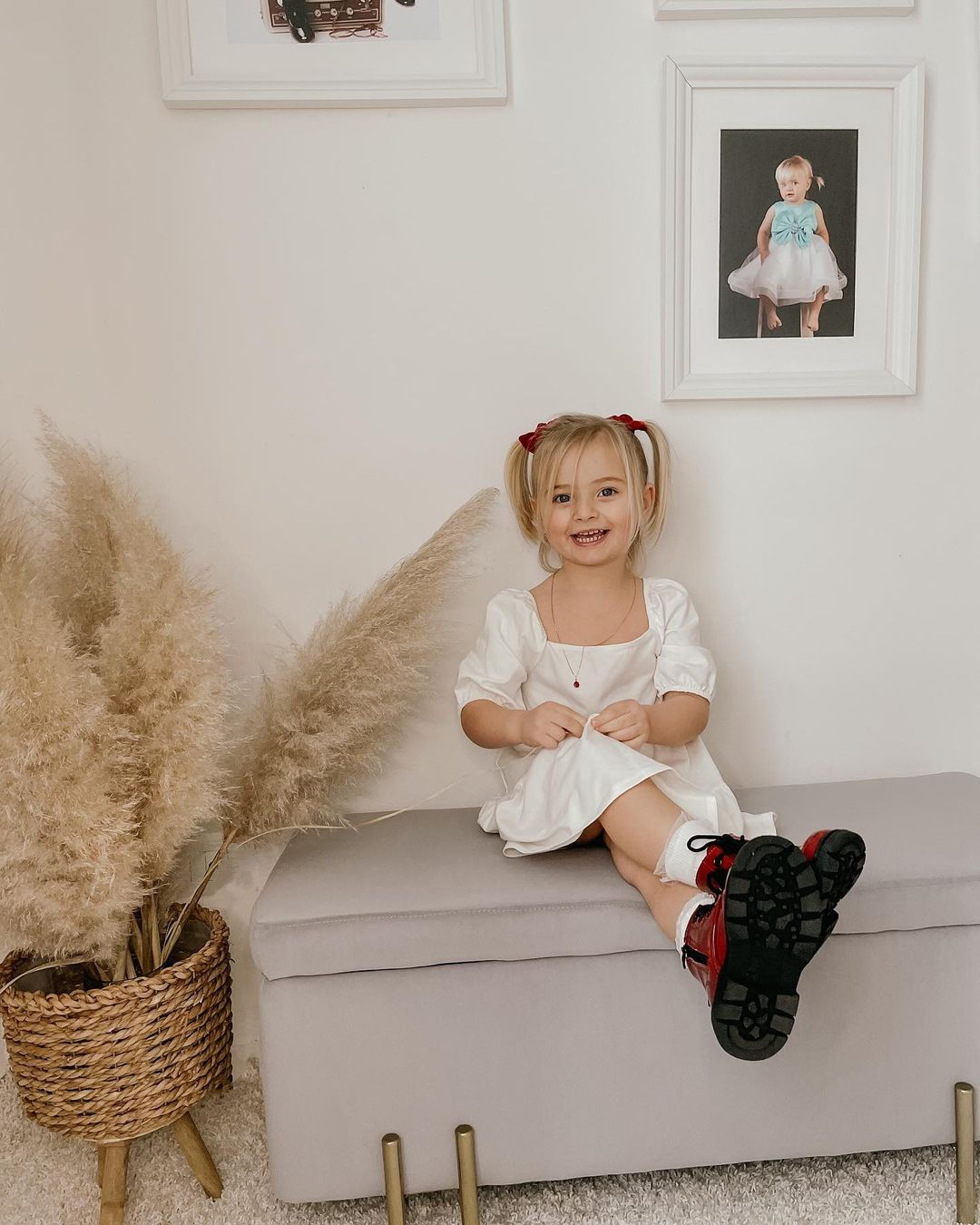 Una bambina in abito bianco è seduta su una panca grigia, sorridente. Ci sono delle foto sul muro.
