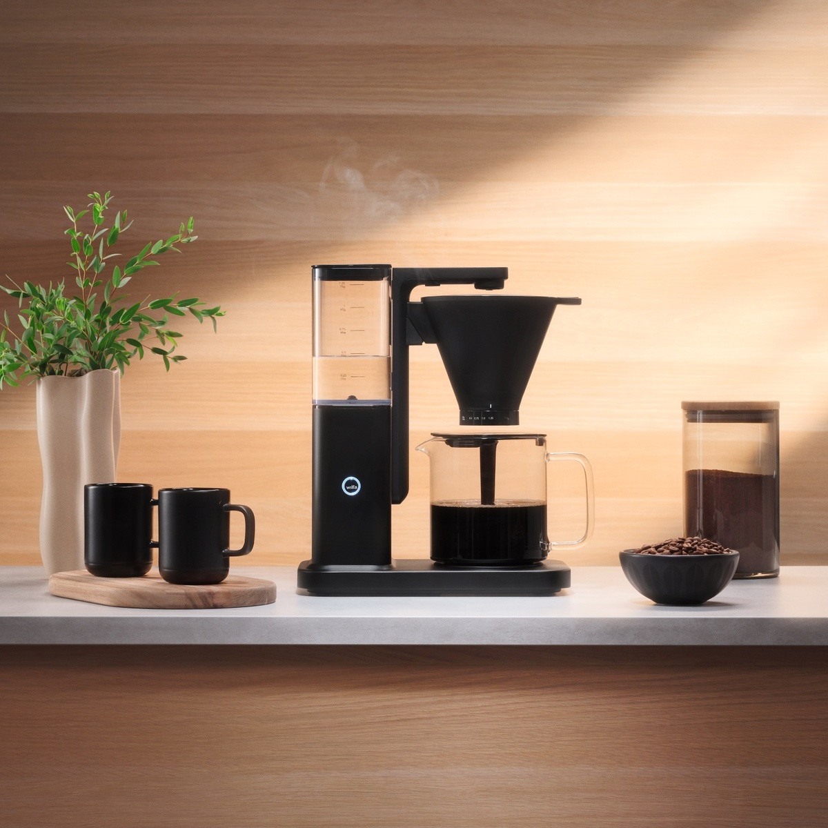 Kaffeemaschine mit schwarzen Tassen, Bohnen und einer Vase mit Grün auf einer weißen Theke, Holz-Hintergrund.